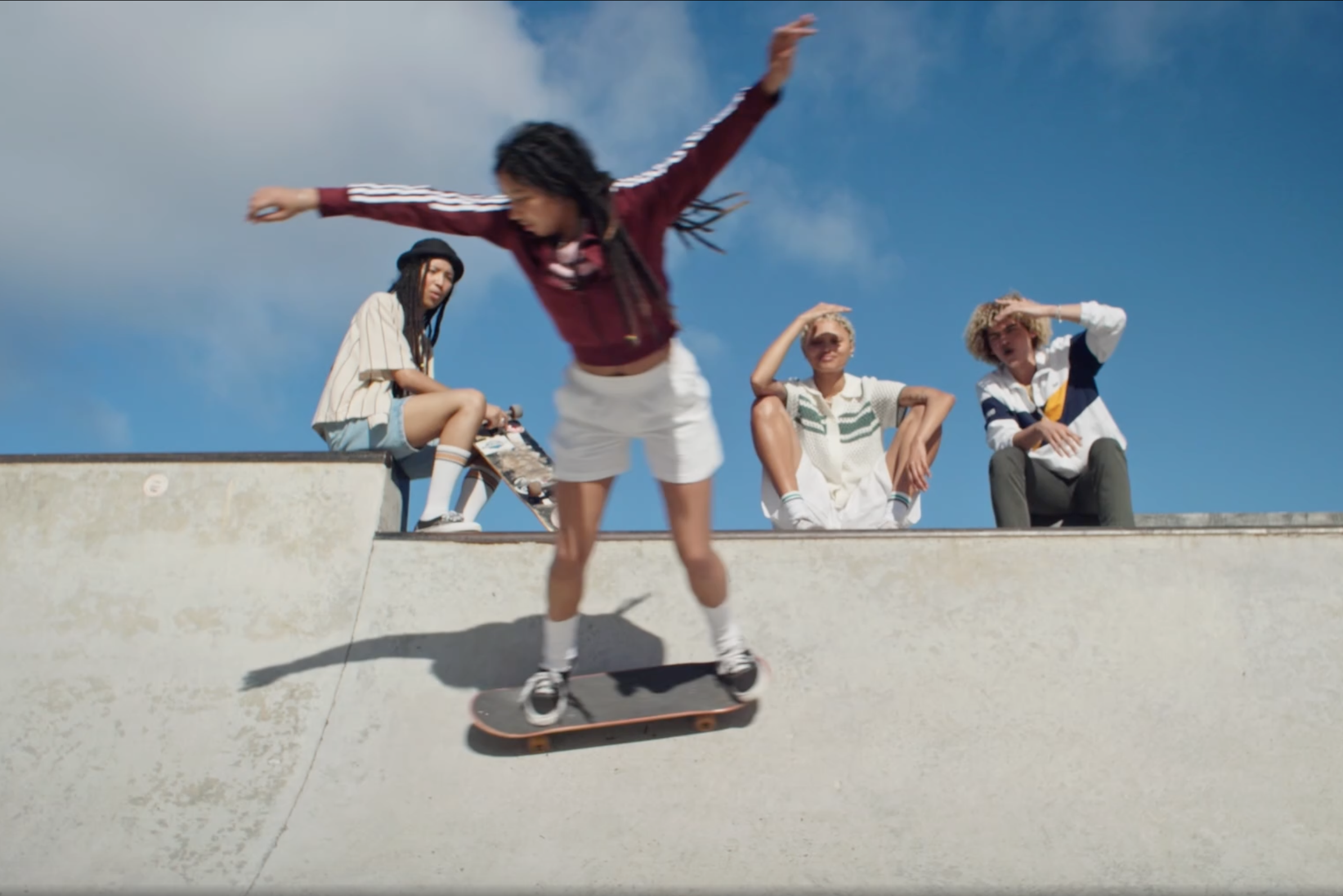 A person skateboarding on a skate ramp while four others sit on the edge watching, with blue sky and clouds in the background.