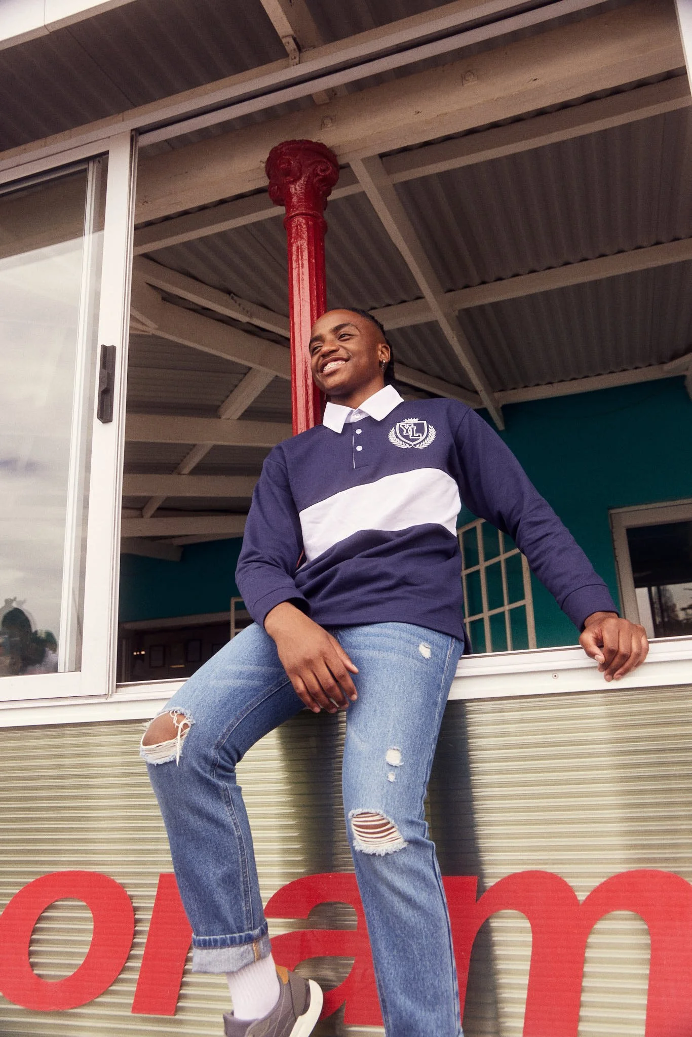 A person of color wearing a navy and white long-sleeve shirt and ripped jeans, sitting on a windowsill outside a building with a corrugated metal roof and a red column behind them, smiling and looking to the side.