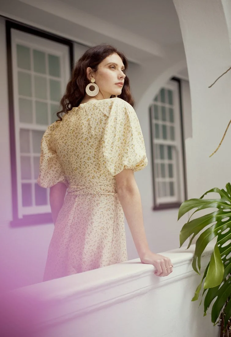 Fashion photography by Justi Munitz - A woman with dark curly hair and large earrings leans on a white balcony railing, looking thoughtfully to the side, next to a green plant indoors near windows.