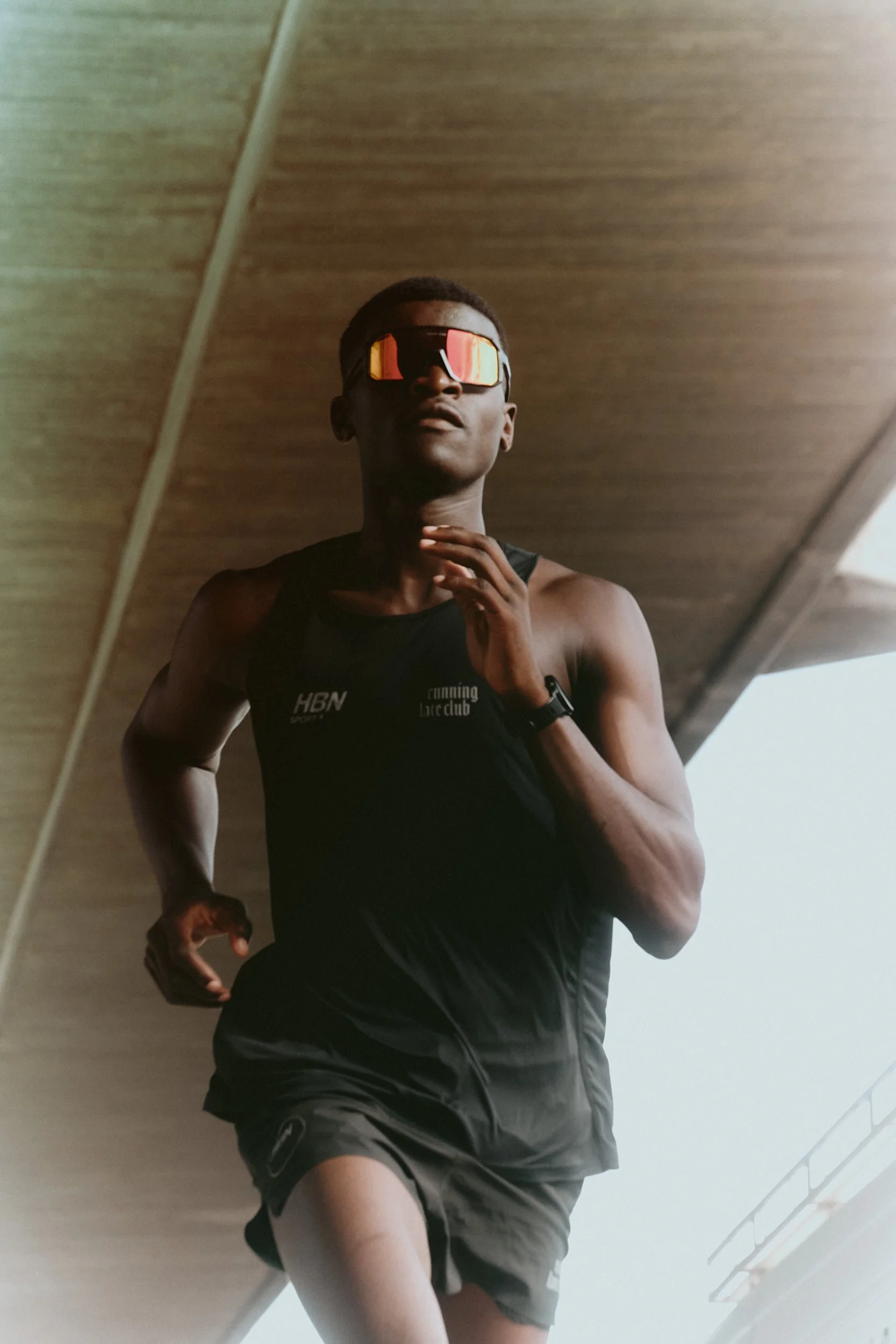 A man running indoors, wearing sunglasses, a black sleeveless athletic shirt, and shorts, captured from a low angle reflection on a mirror.