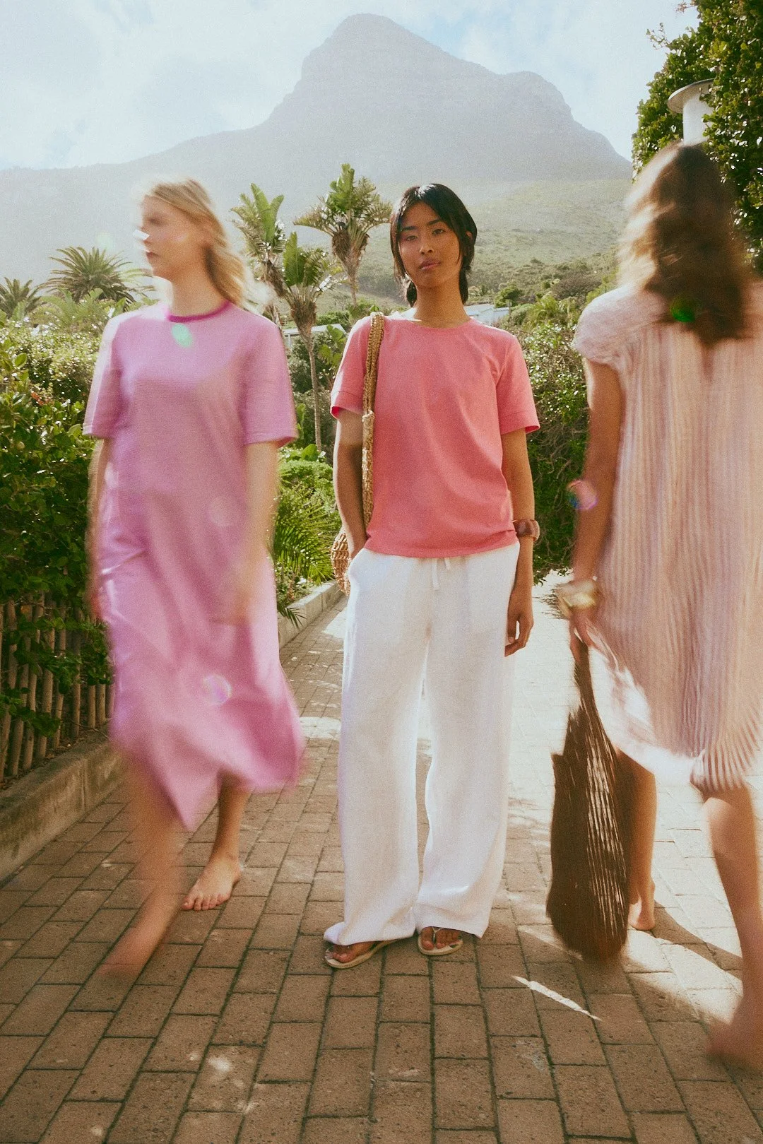 Three women walking outdoors on a brick path, with mountains and greenery in the background. The woman in the center wears a pink T-shirt and white pants, while the other two women wear pink dresses and carry bags.