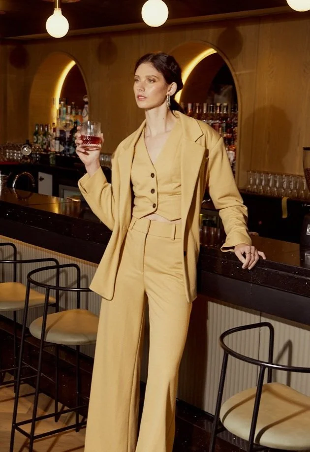 Fashion Photography by Justin Munitz - A woman in a yellow suit holding a glass of red wine standing at a bar inside a restaurant or bar with a warm ambiance.