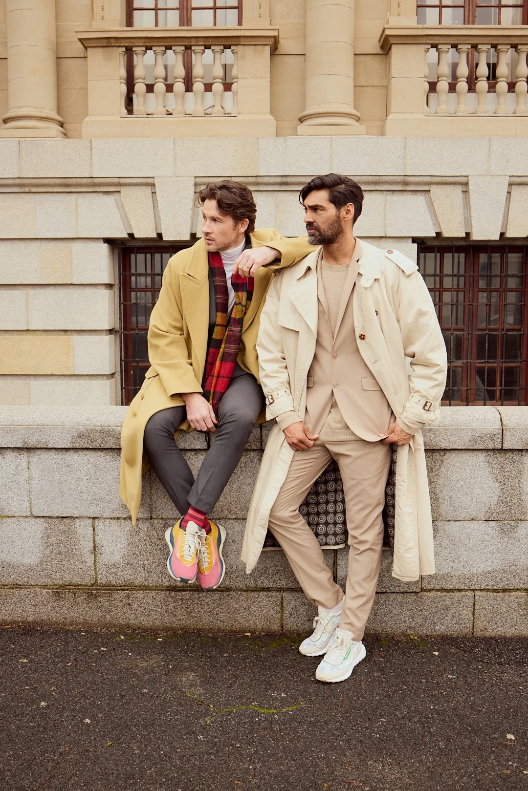 Two men dressed in beige and yellow trench coats sitting and standing on a stone wall in front of a beige building with columns and fence windows.