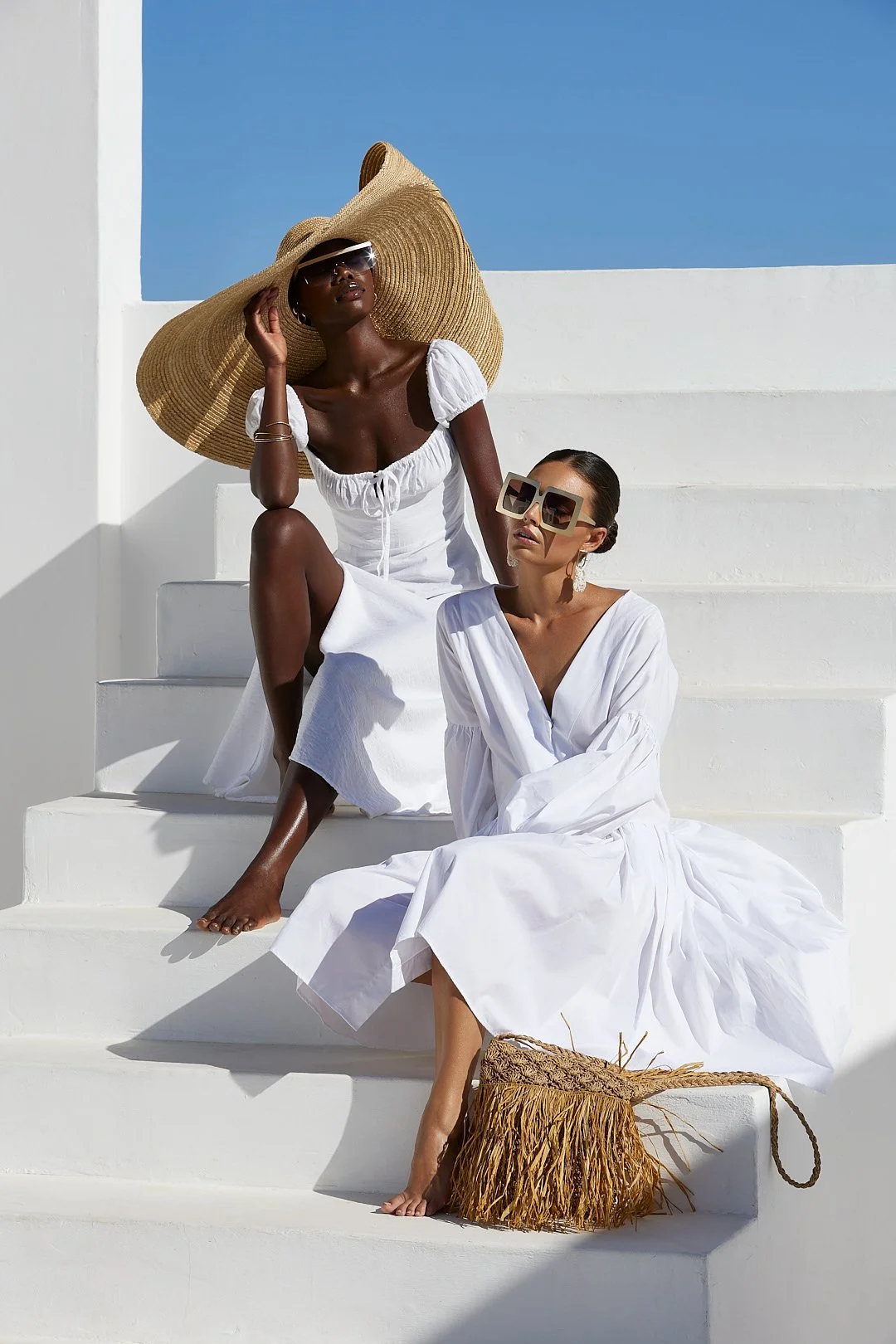 Fashion photography by Justin Munitz . Two women dressed in white sitting on white steps outdoors, with one wearing a large sunhat and the other wearing oversized sunglasses, against a backdrop of a blue sky.