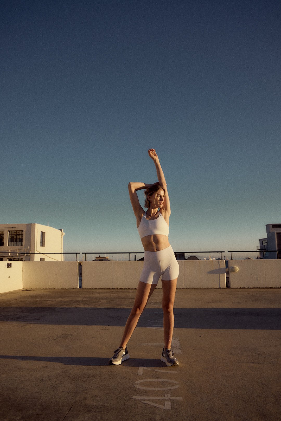 A woman in workout clothes stretching on a rooftop parking lot at sunset.