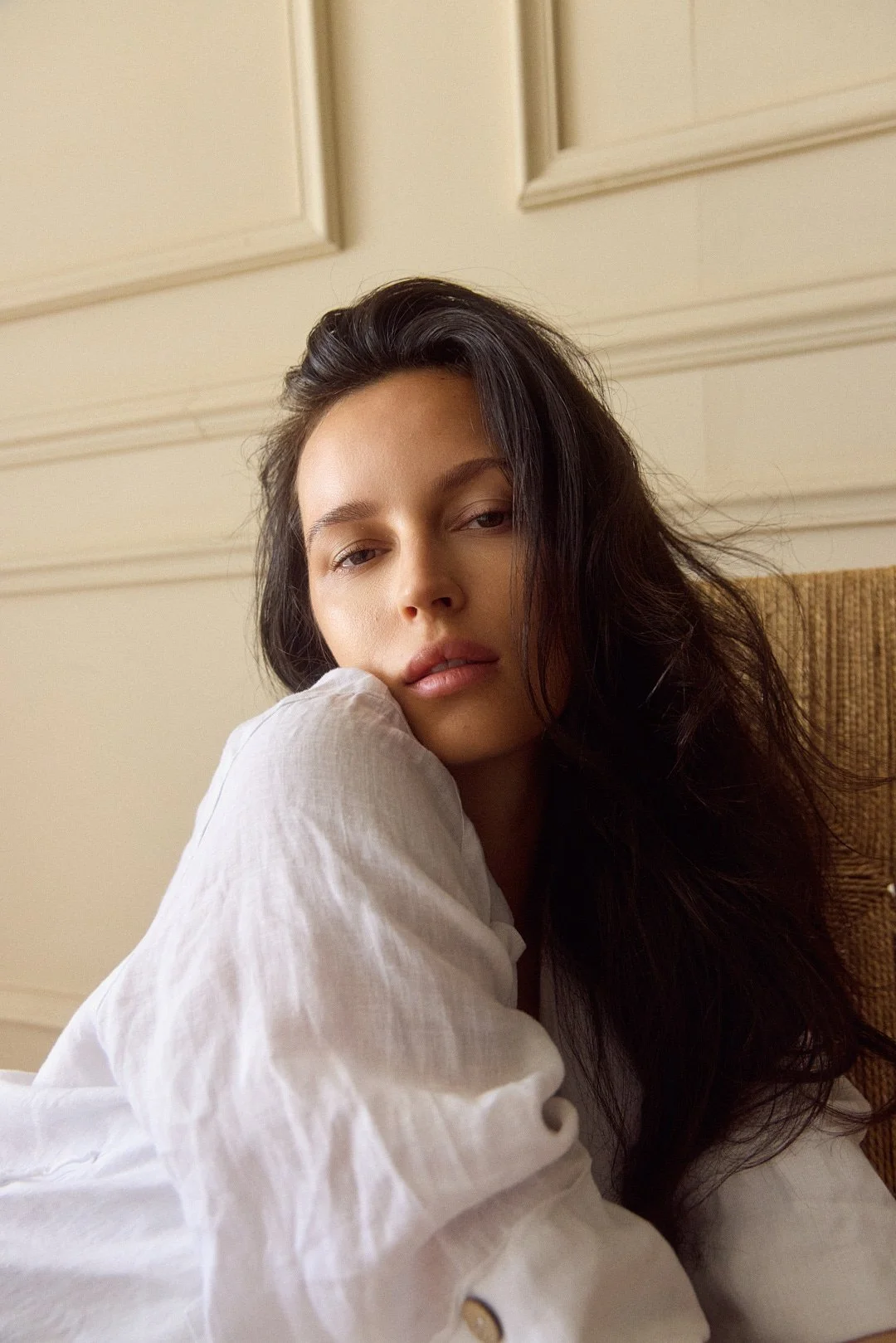 A young woman with long dark hair resting her head on her arm, looking at the camera, wearing a white shirt, in a beige room.