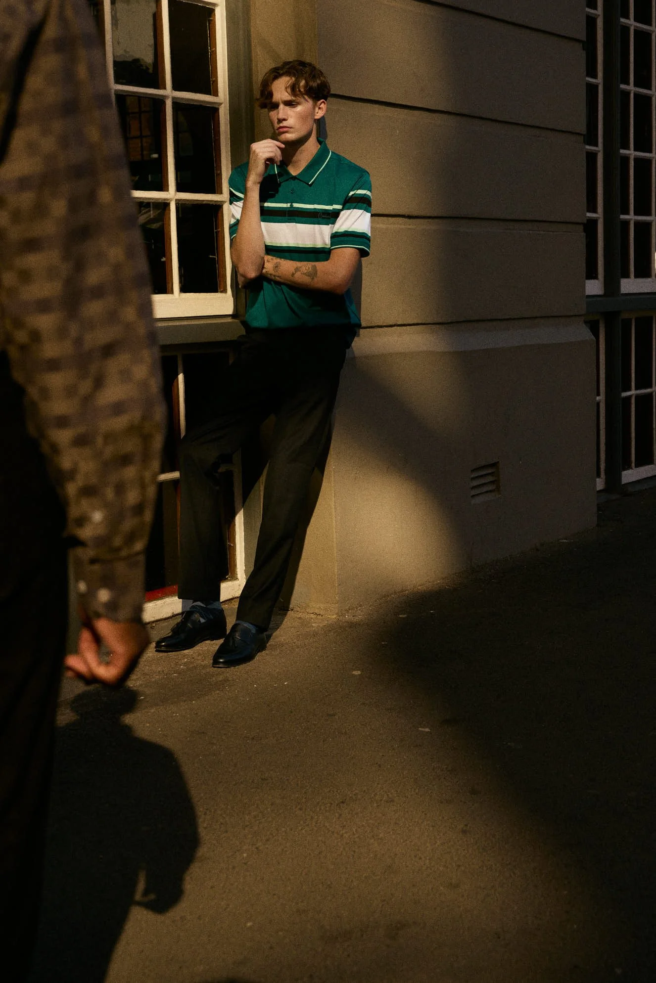 A young man wearing a teal and white striped polo shirt and black pants is standing outside on a dimly lit street, leaning against a wall with his arms crossed, while someone in the foreground appears to be in motion.