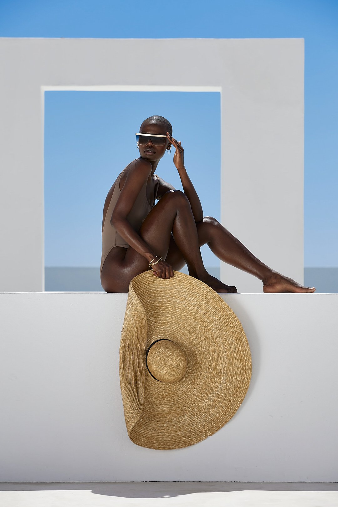 Fashion photography by Justin Munitz — model wearing a beige swimsuit with a straw hat, seated against a white architectural structure and blue sky