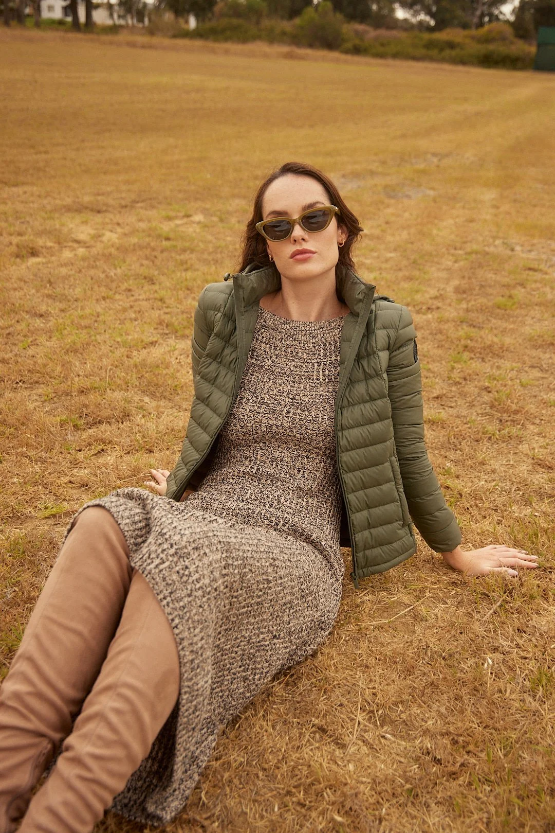 A woman with dark hair, wearing sunglasses, a brown knit dress, an olive green puffer jacket, and tan knee-high boots, sitting on dry grass in an open field.