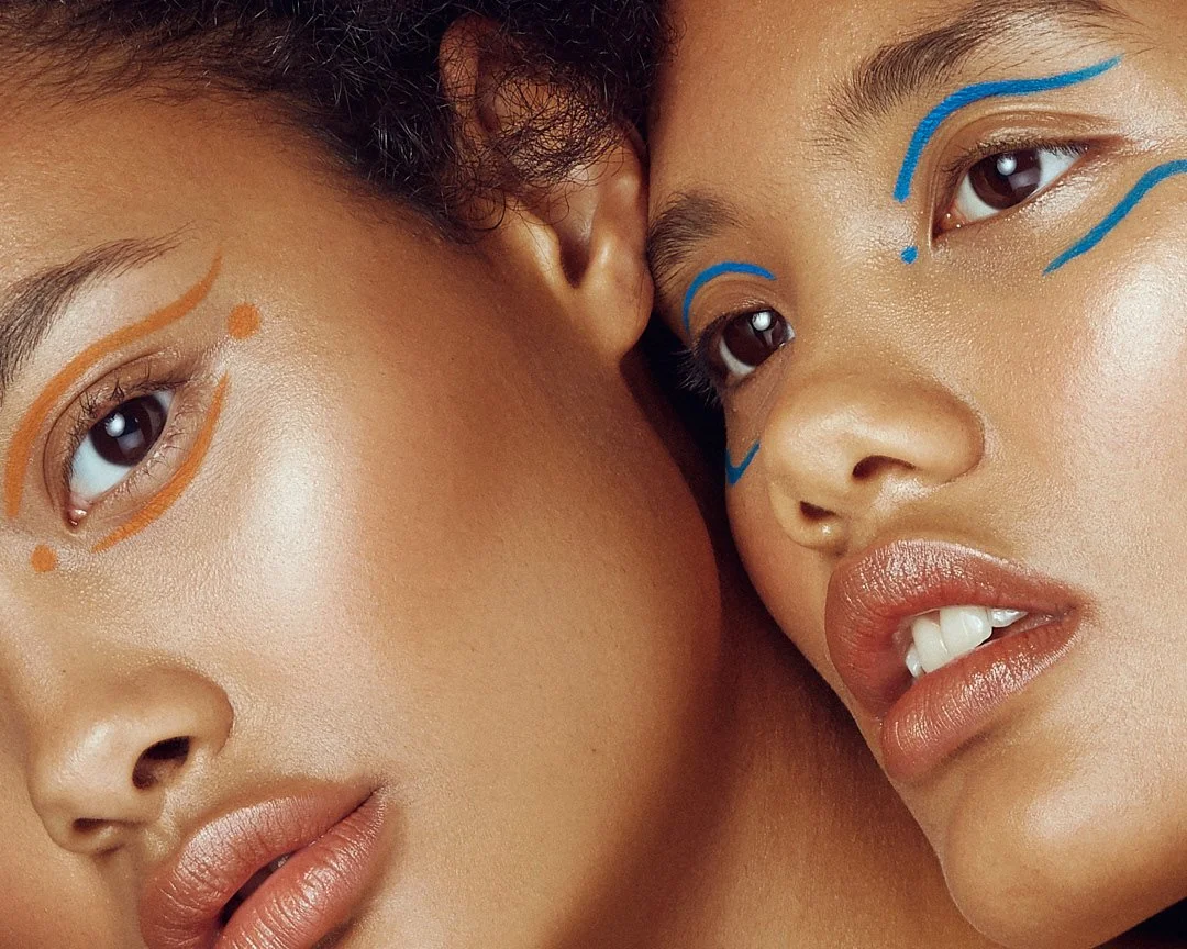 Close-up beauty portrait of two models with colourful face paint, one orange and one blue, looking at each other, photography by Justin Munitz.