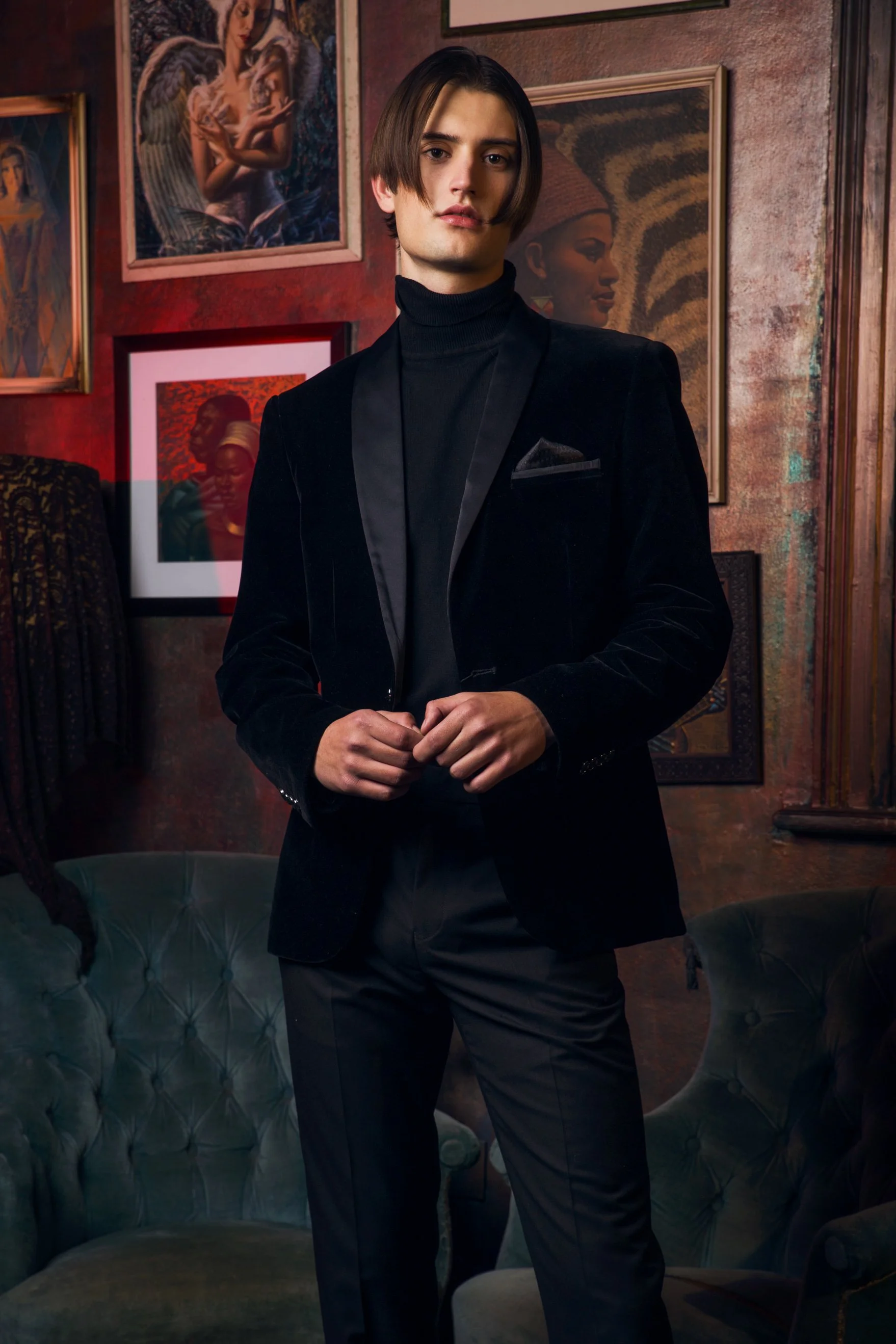 Fashion photography by Justin Munitz — model wearing a black turtleneck and velvet blazer, standing in a dimly lit room.