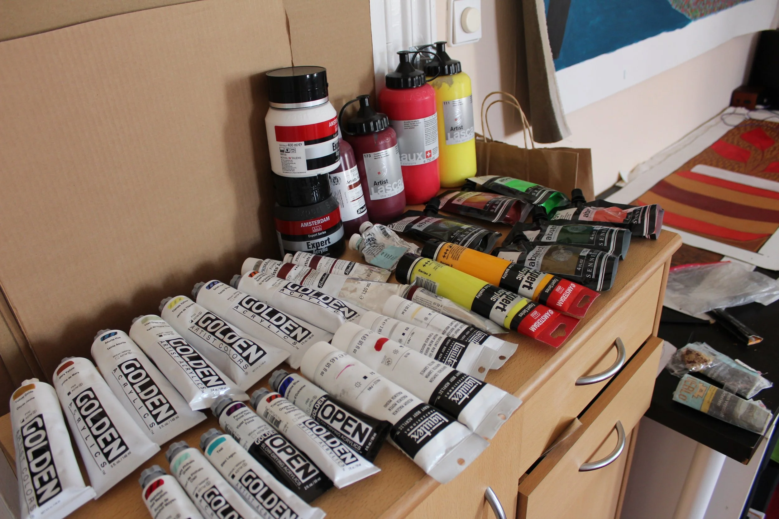 A wooden dresser with various tubes of acrylic paint, bottles of liquid paint, and paint containers arranged on top. In the background, there is a paper bag and a colorful painting leaning against the wall.