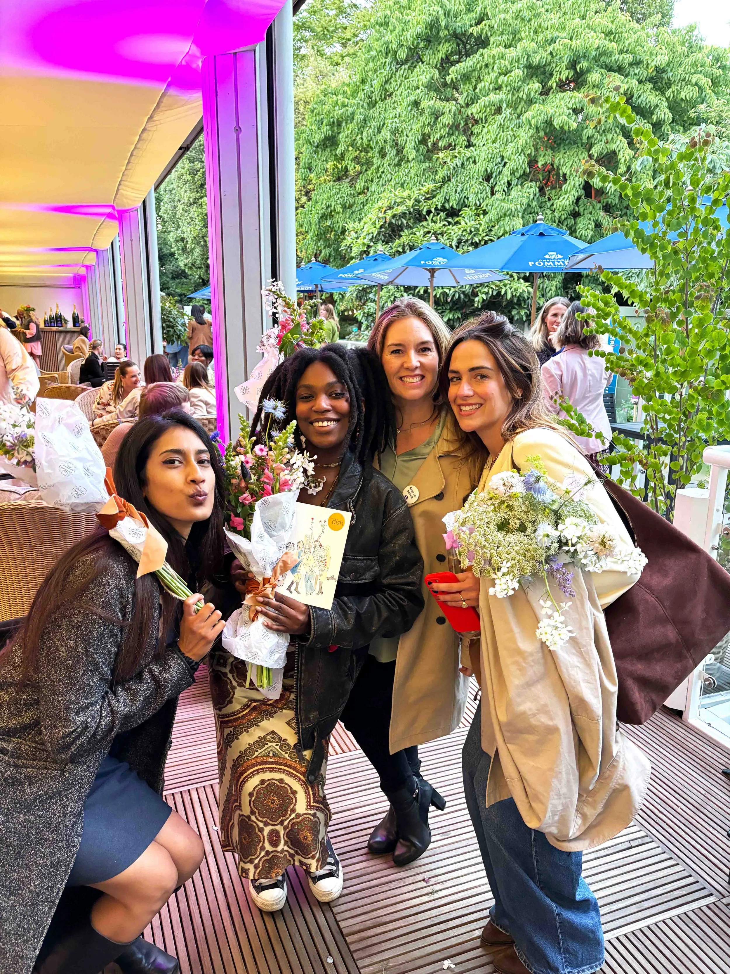 Four women standing together at a social gathering, holding bouquets of flowers, smiling, with a background of other people, outdoor umbrellas and greenery.