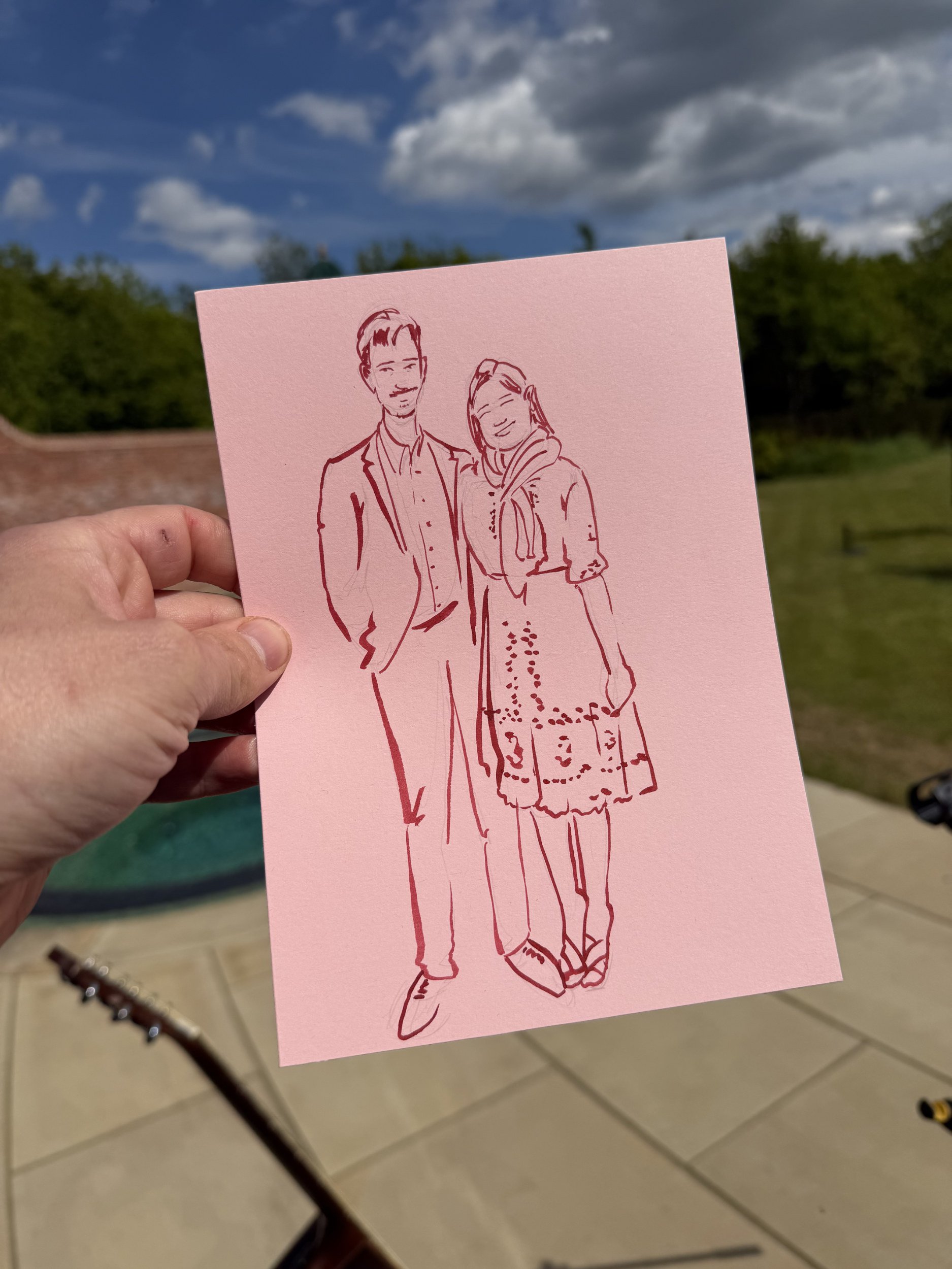 A hand holding a pink card with a line drawing of a man in a suit and a woman in a dress standing close together, outdoors with a cloudy sky and greenery in the background.