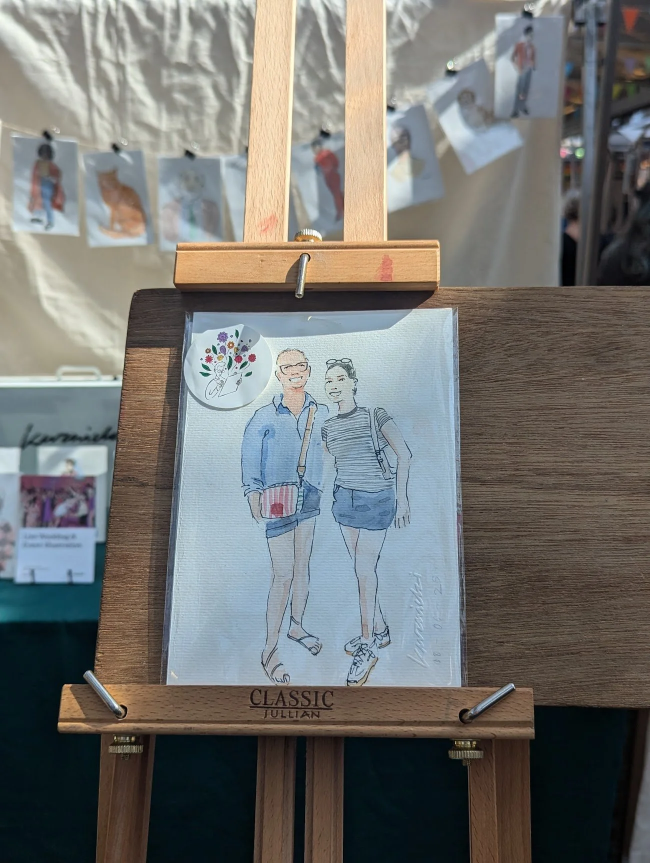 A framed watercolor illustration of a smiling man and woman standing outdoors, with people and various artworks visible in the background.