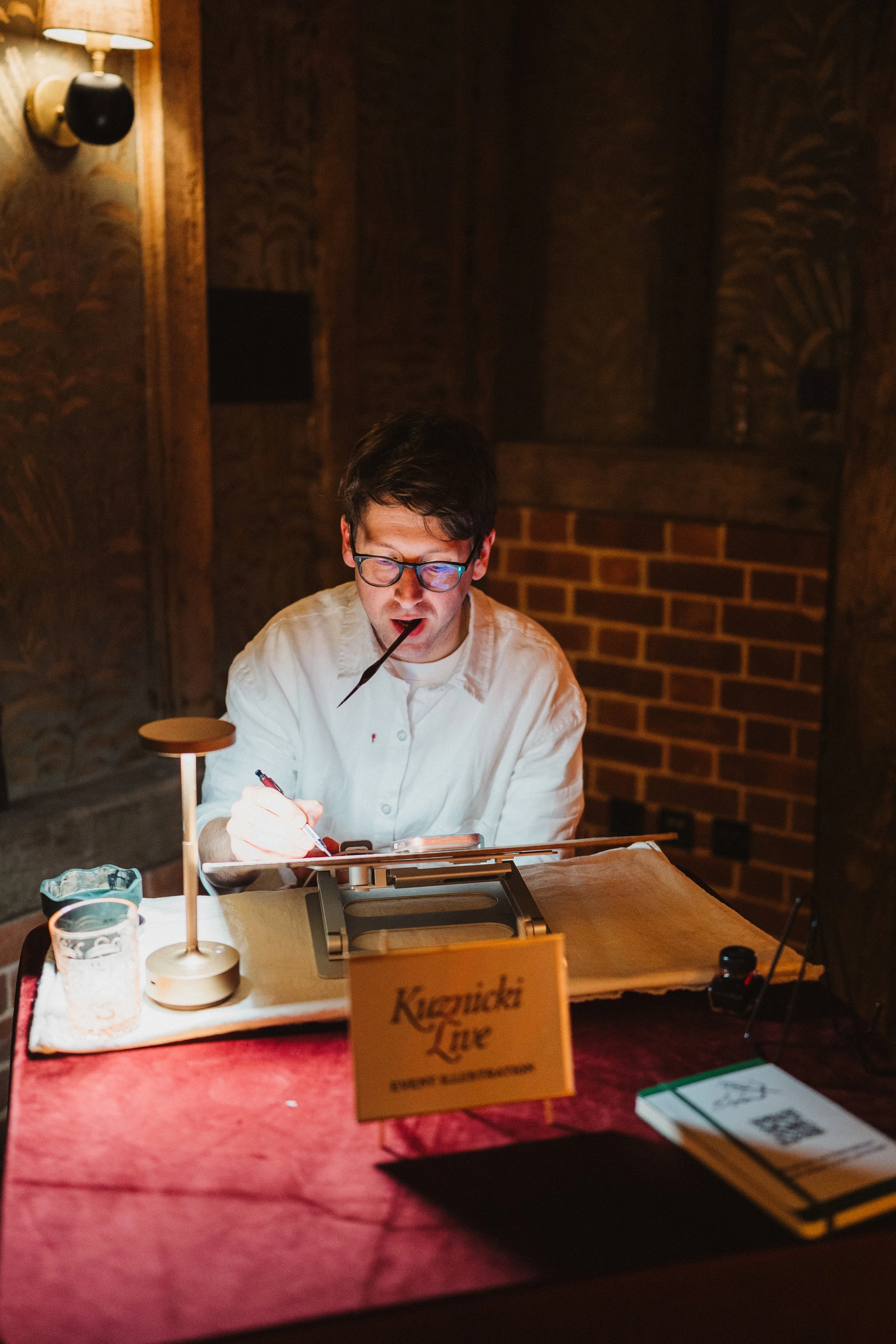 A man with glasses and dark hair sits at a table, writing on a clipboard in a dimly lit room with wooden walls and a brick fireplace background. There are various items on the table, including a small lamp, a glass, and a sign that reads 'Kazuki's Life'.