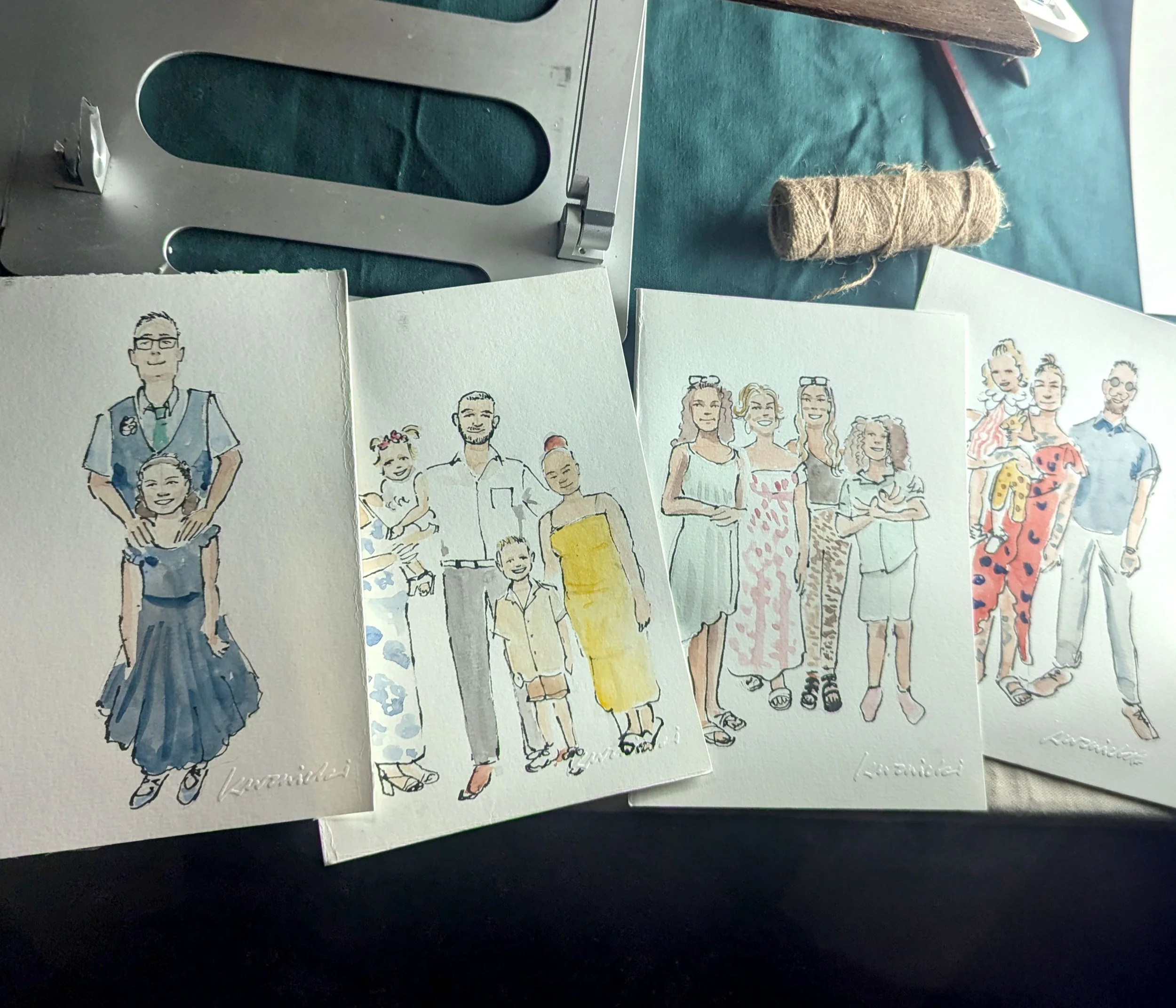 Watercolor illustrations of various families, each with different groupings of adults and children, arranged on a work surface with art supplies and a spool of twine in the background.