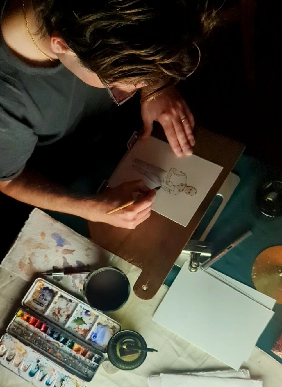 Person drawing a cartoon figure at a desk with watercolor paints, paintbrush, and paper.