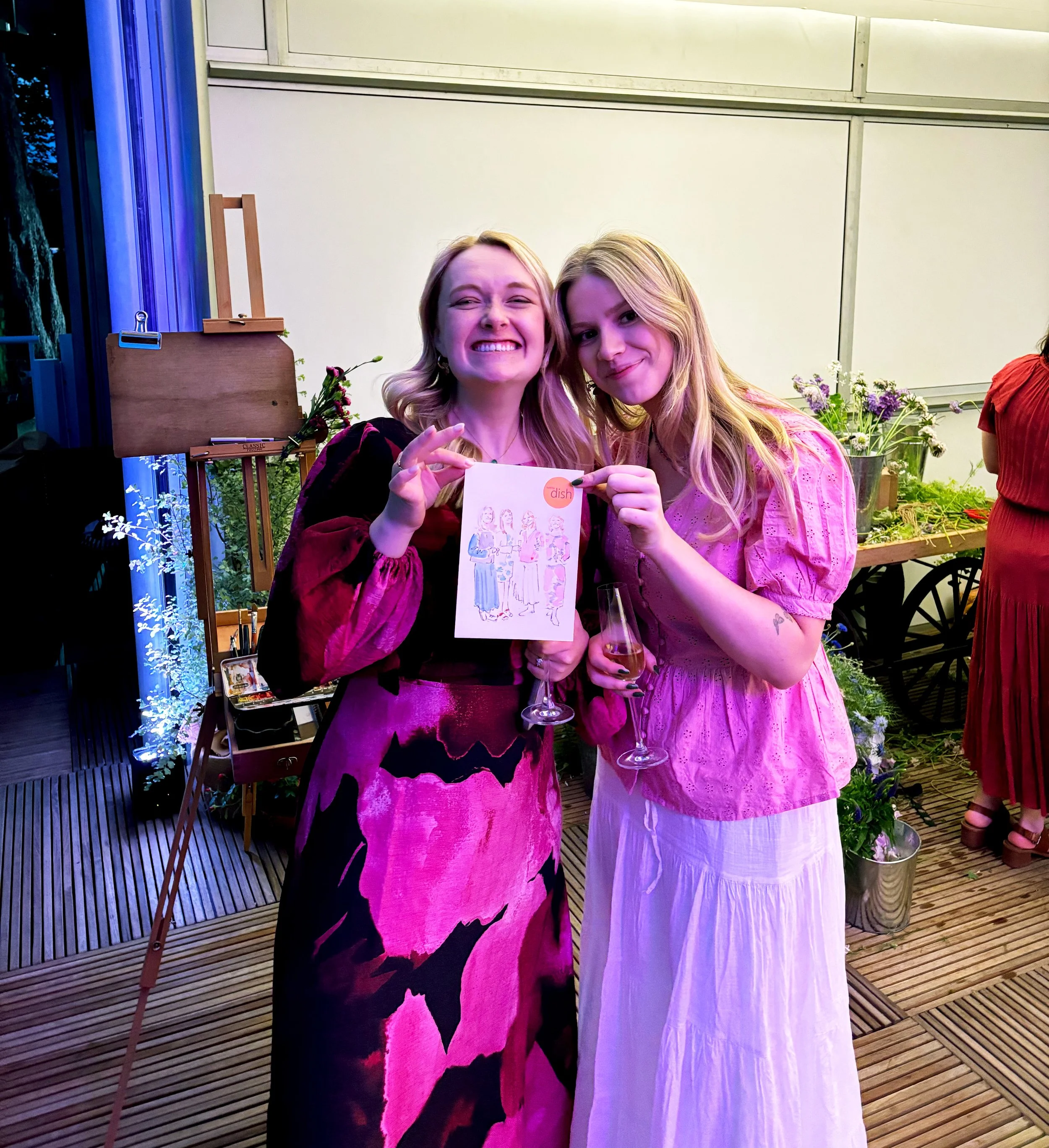 Two young women smiling and holding drinks and artwork at a social event, standing on a wooden deck with plants and a table of flowers in the background.