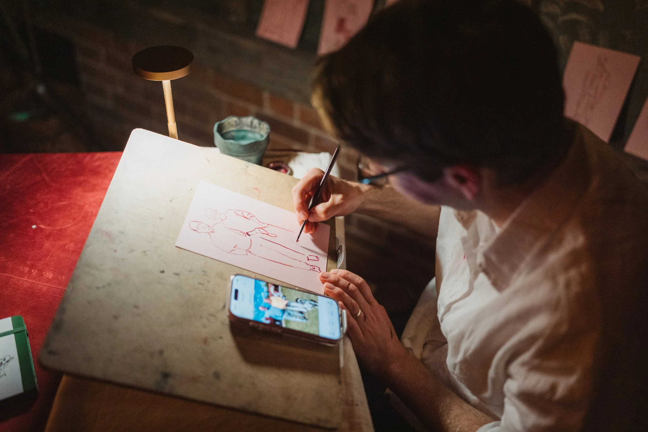 Person drawing a picture of two figures on a white piece of paper at a wooden table, with a smartphone showing a photo of a scene with people, a small table lamp, and a glass of water nearby.