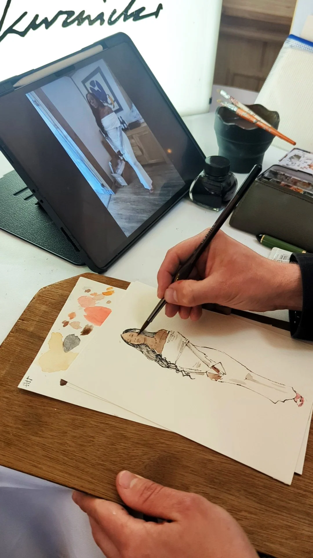 An artist's hand painting a fashion sketch of a woman in a long dress, with a palette of watercolors on a wooden board, a tablet displaying a reference photo of a woman in white pants and a top, and art supplies on a white table.