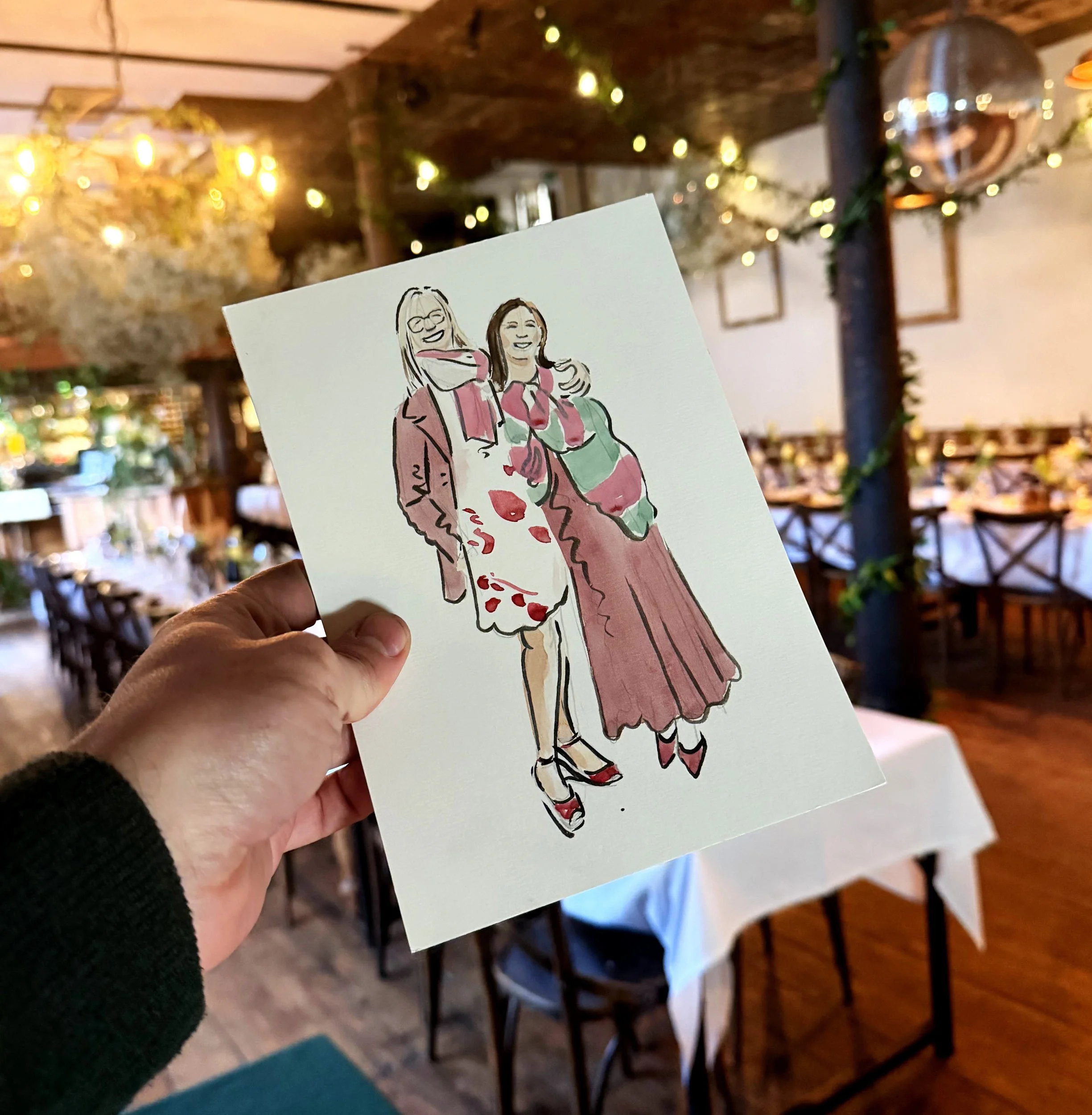 A hand holding a cartoon illustration of two women smiling and posing together inside a cozy, decorated restaurant or cafe with warm lighting and wooden furniture.