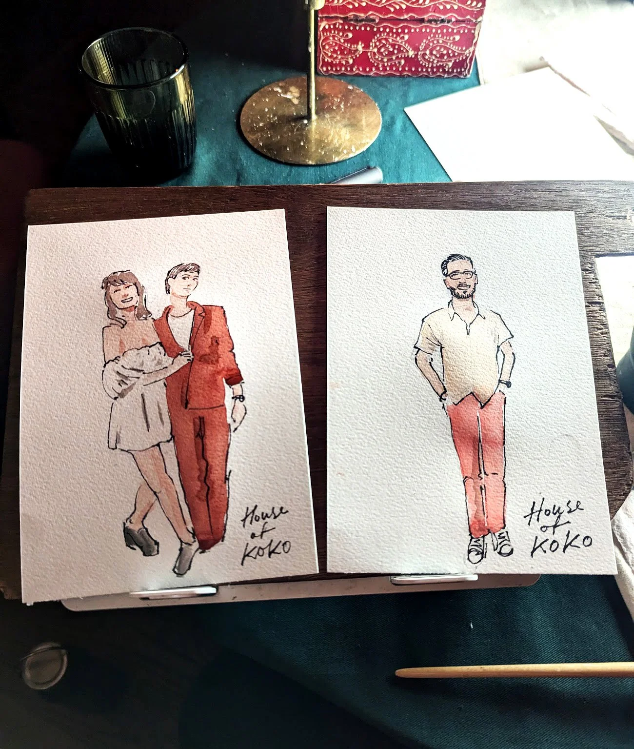 Two watercolor sketches on paper. The left sketch depicts two women standing close, smiling, with one putting an arm around the other, wearing casual and stylish outfits. The right sketch shows a man with glasses and a beard, standing with hands in p