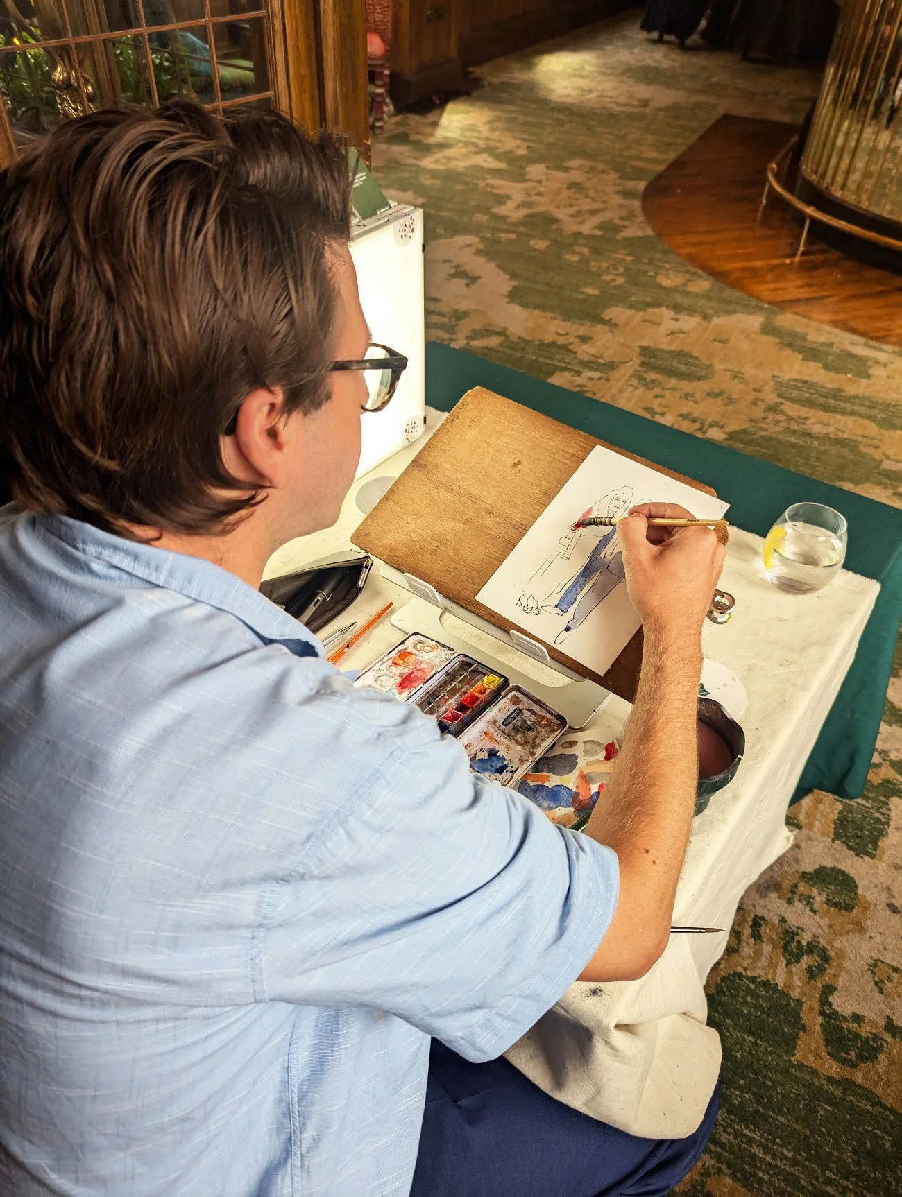 A man with glasses, wearing a light blue shirt, is sitting at a table and painting a fashion illustration of a woman in a red top and blue pants. The table has watercolor supplies, a glass of water with lemon, and a paper towel.