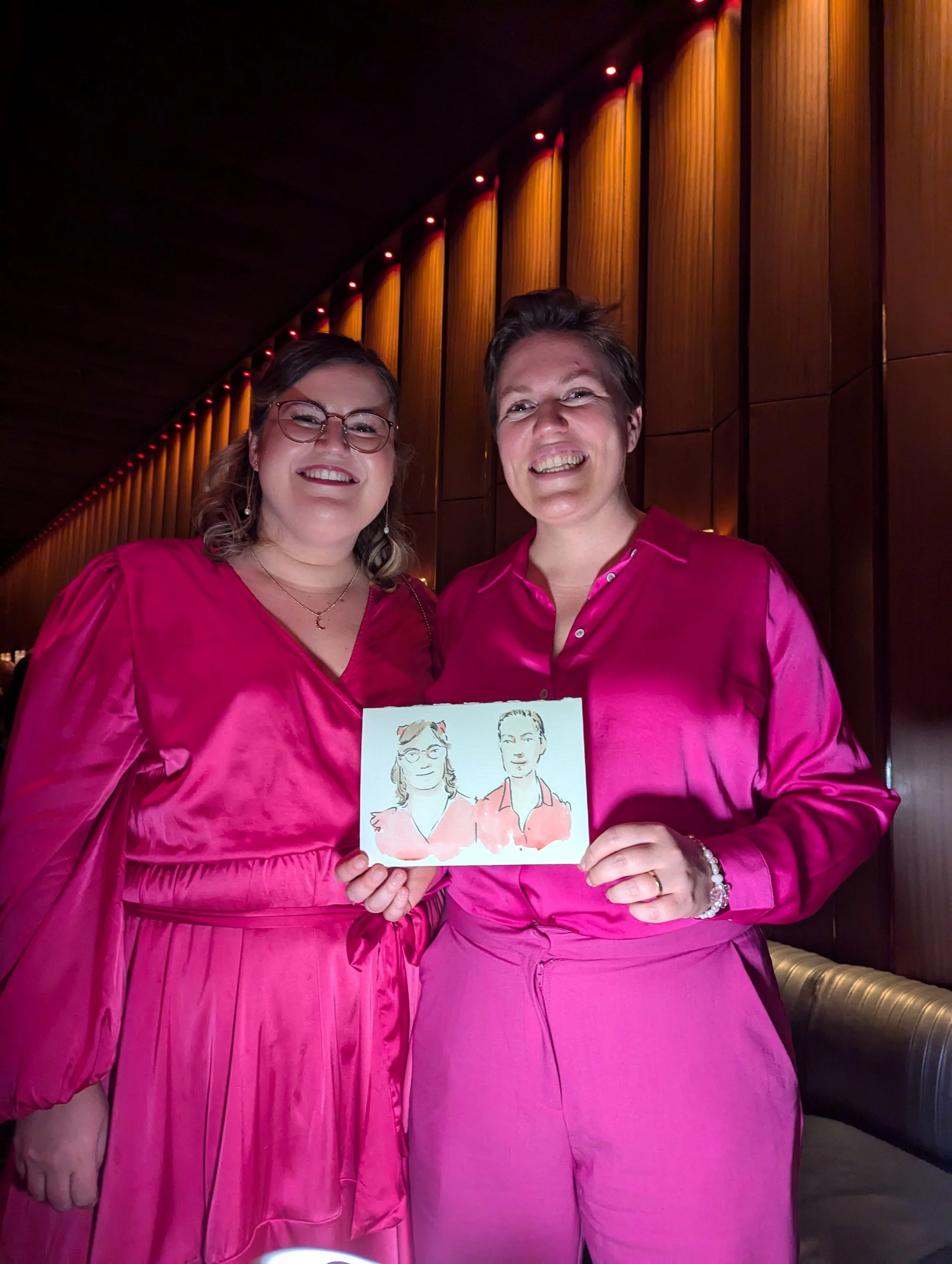 Two women in pink satin dresses smiling and holding a drawing of themselves in front of a dark wooden wall with red lighting.