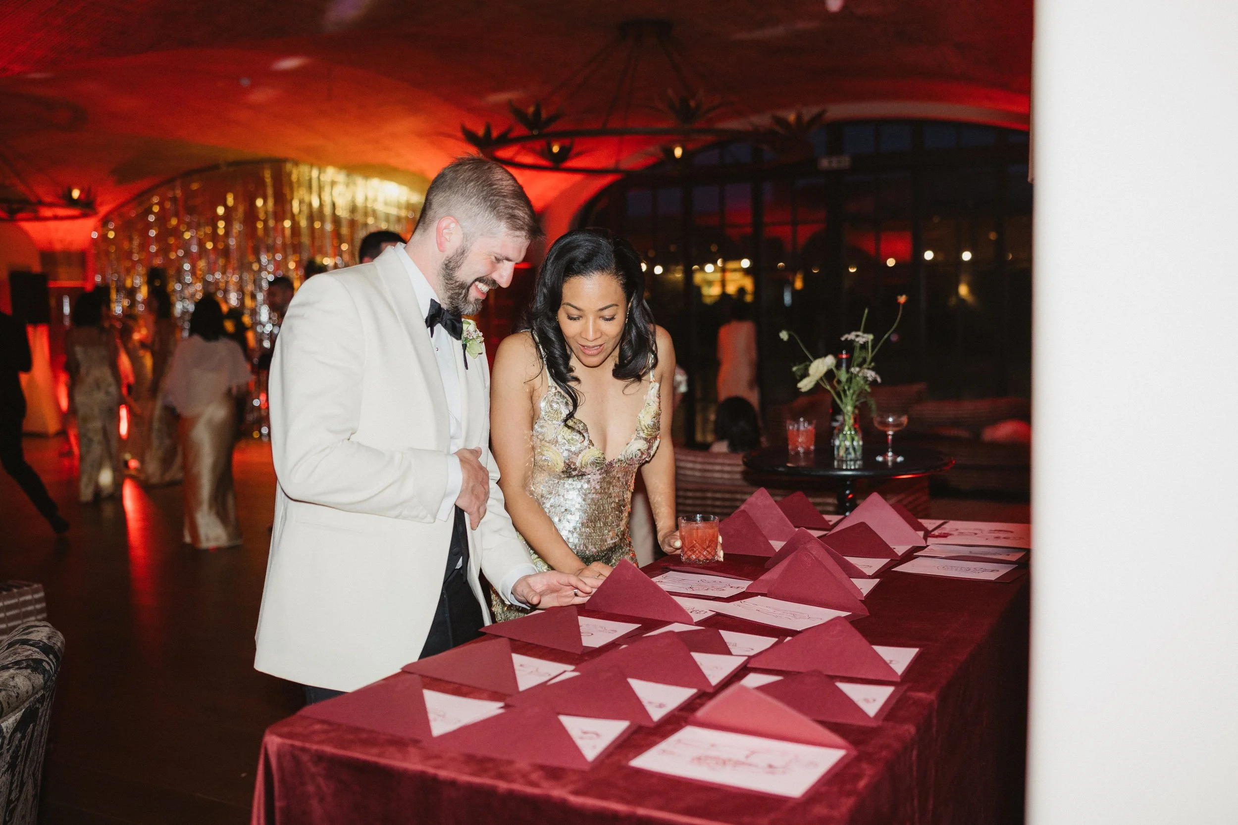 A wedding reception with a bride and groom in formal attire gathering wedding programs or place cards on a table covered with a red tablecloth in a dimly lit venue with warm lighting and hanging decorations.