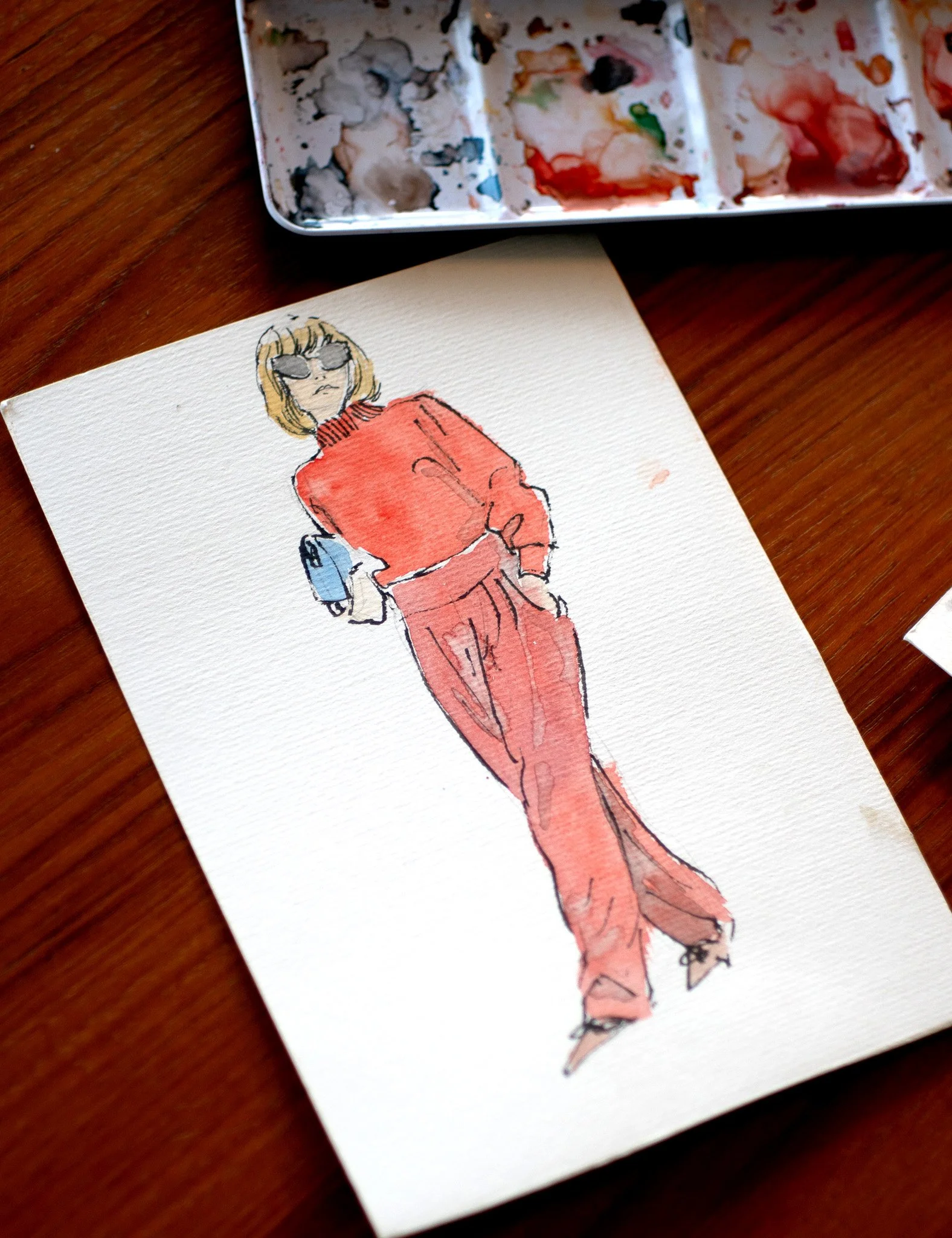 A watercolor illustration of a woman with blonde hair wearing sunglasses, a red jacket, and red pants, standing with her hands in her pockets on a textured white paper.
