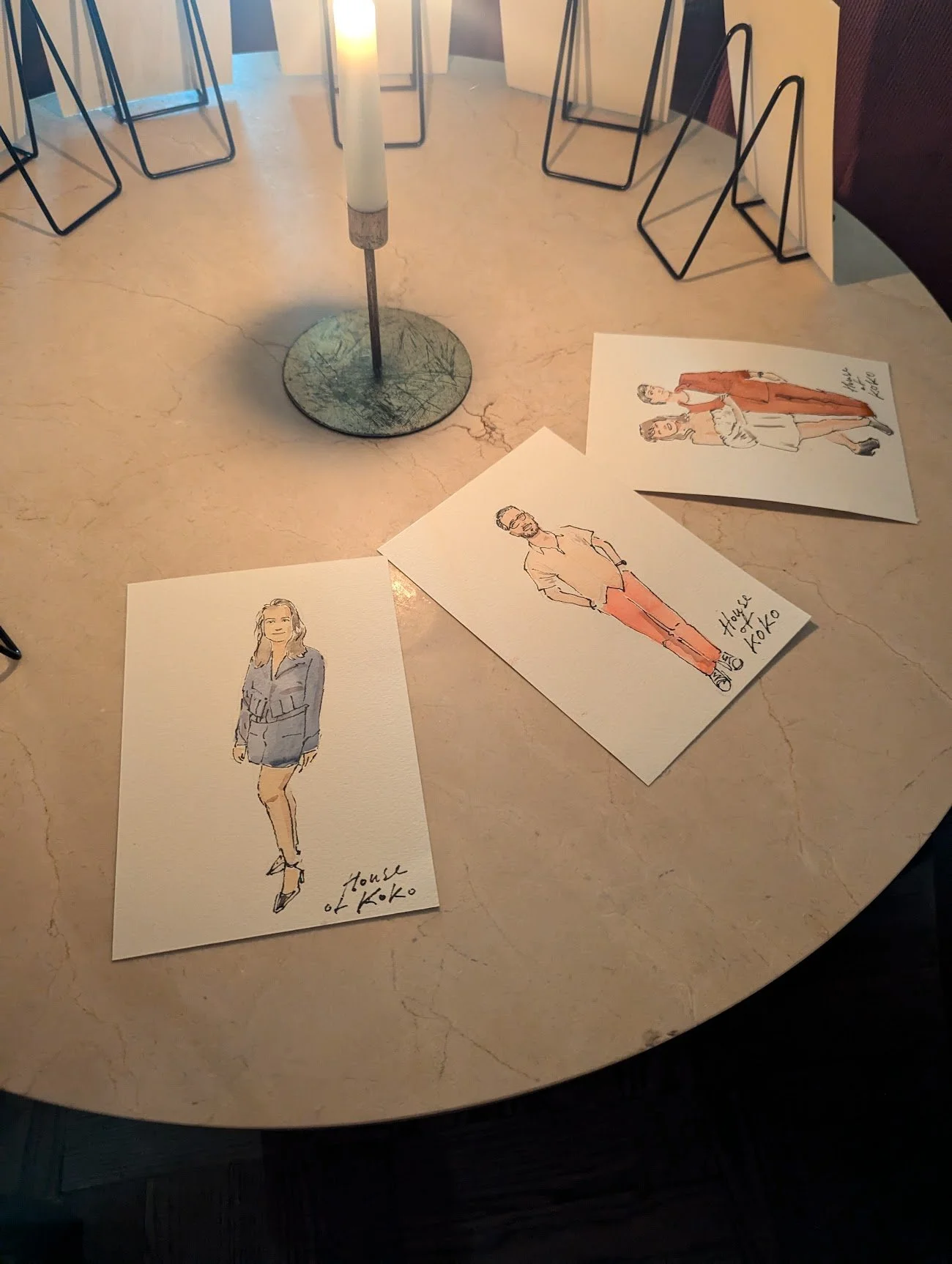Watercolor illustrations of three women and one man on separate sheets of paper, placed on a light-colored table, with a candle and some black metal chair legs in the background.