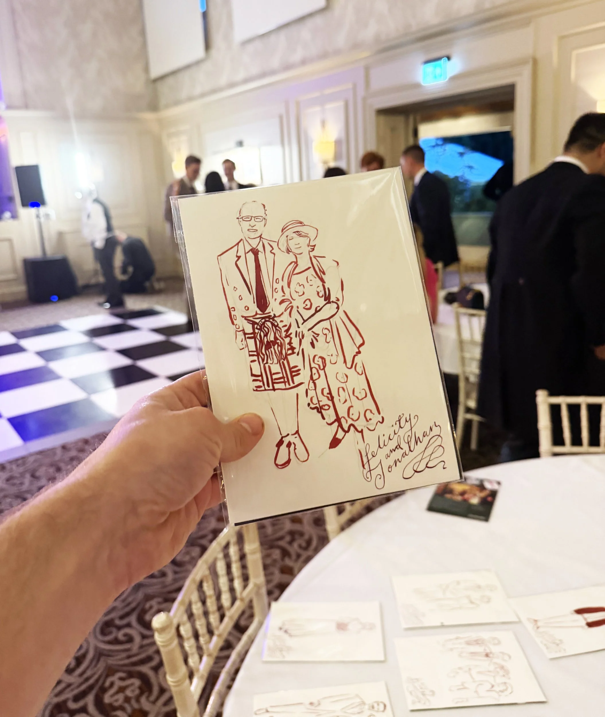 A person holding a greeting card with a line drawing of a man in a suit and a woman in a dress, in an event hall with people socializing in the background and displays of similar cards on a table.
