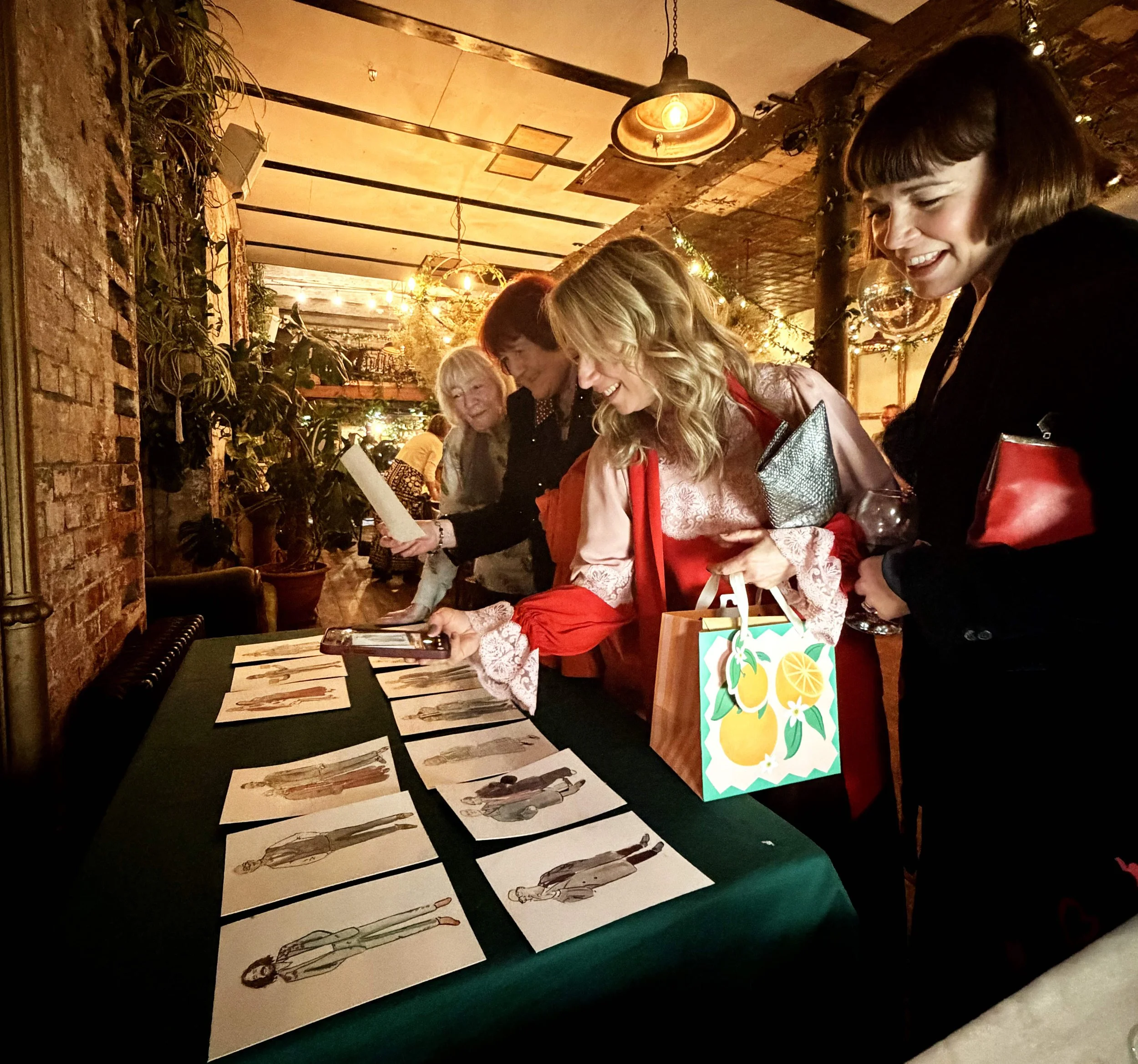 People at a table looking at fashion illustrations and a decorated gift bag, in a warm, cozy restaurant or cafe with hanging lights and plants.
