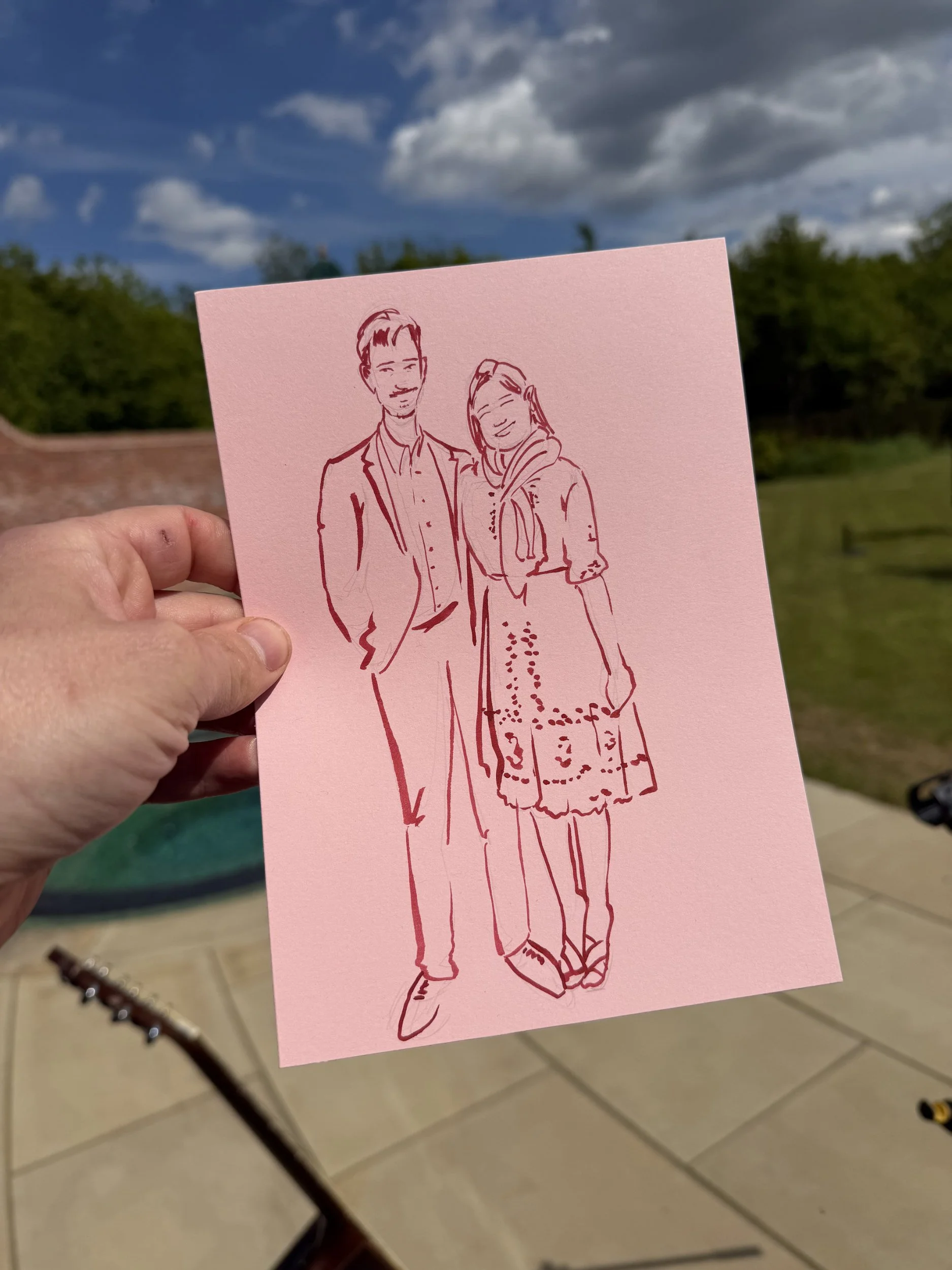 Hand holding a pink card with a line drawing of a man and woman smiling, outdoors with trees and cloudy sky in the background.