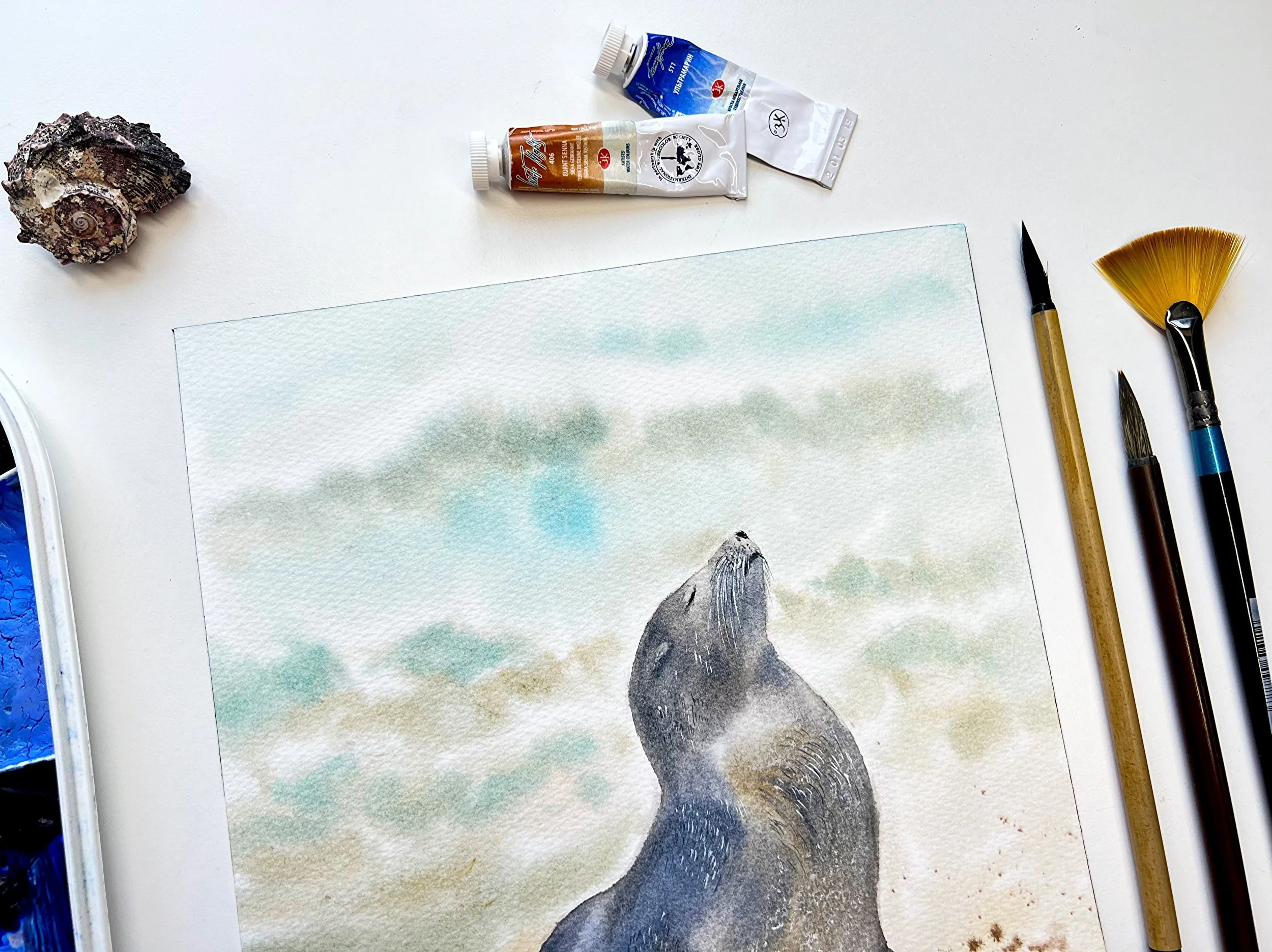 Close up of an original watercolor features a gentle Sunbathing Seal on the beach, painted in impressionistic style. in a setting with the original art supplies