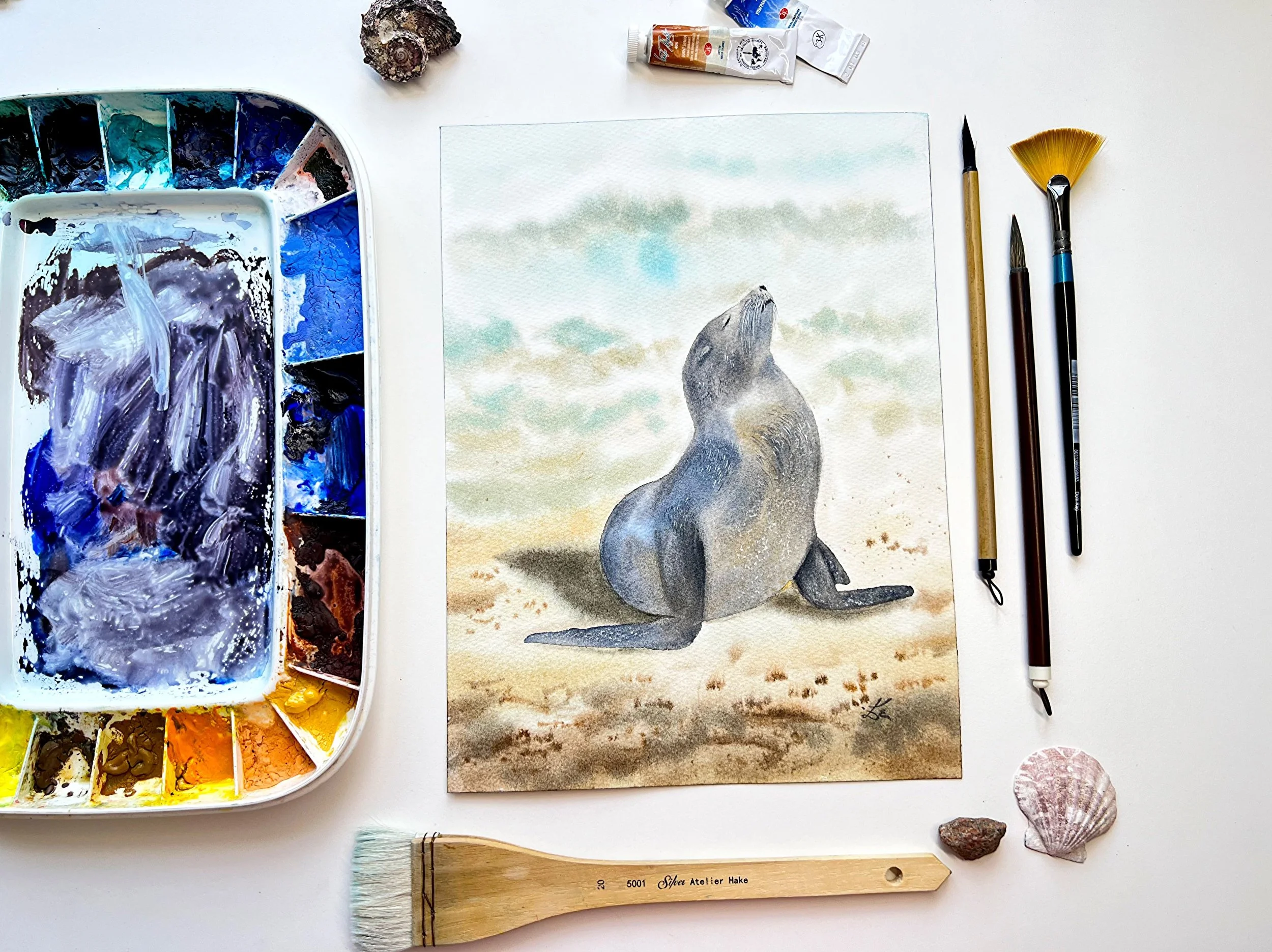 Close up of an original watercolor features a gentle Sunbathing Seal on the beach, painted in impressionistic style. in a setting with the original art supplies