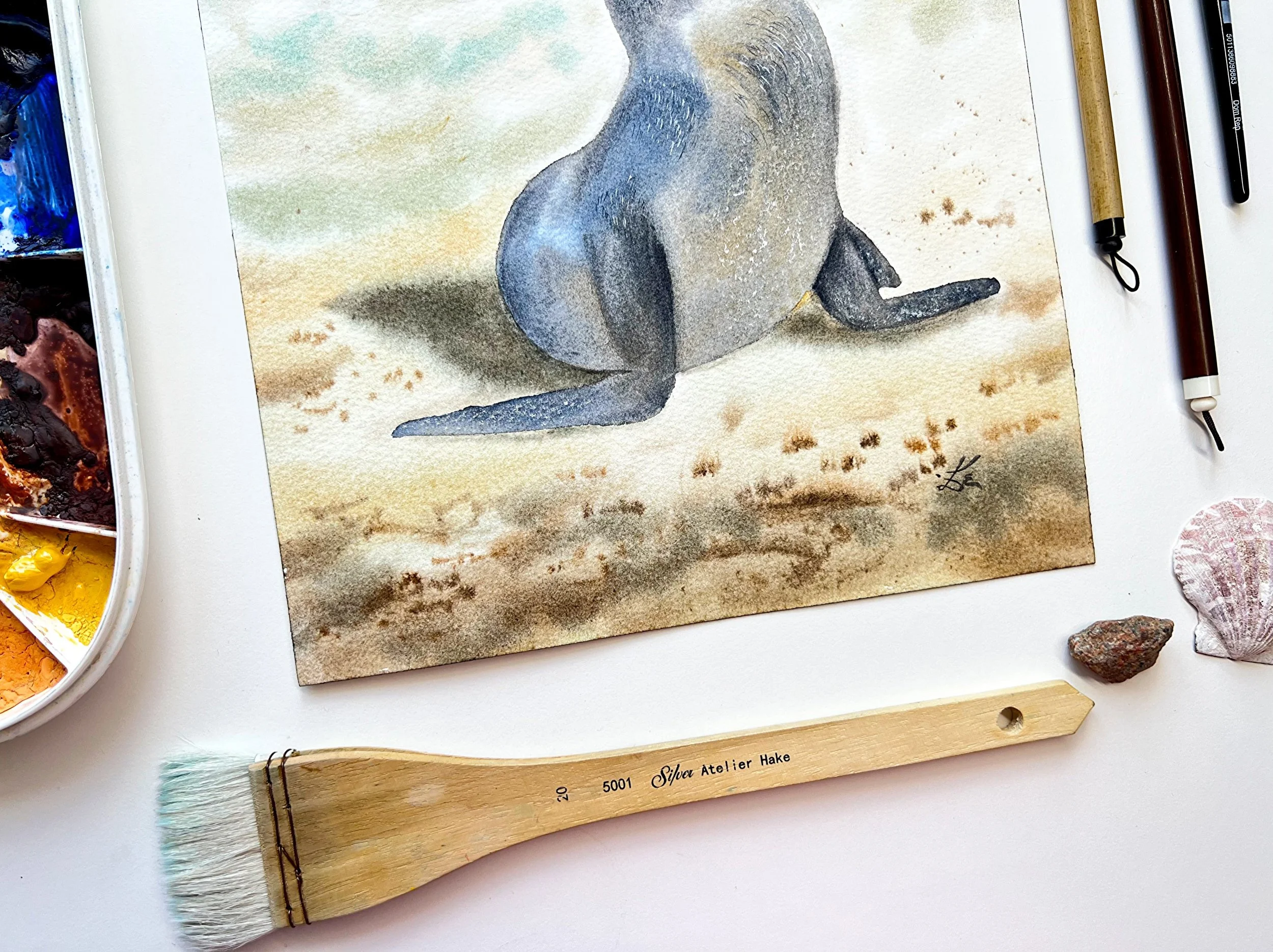 Close up of an original watercolor features a gentle Sunbathing Seal on the beach, painted in impressionistic style. in a setting with the original art supplies