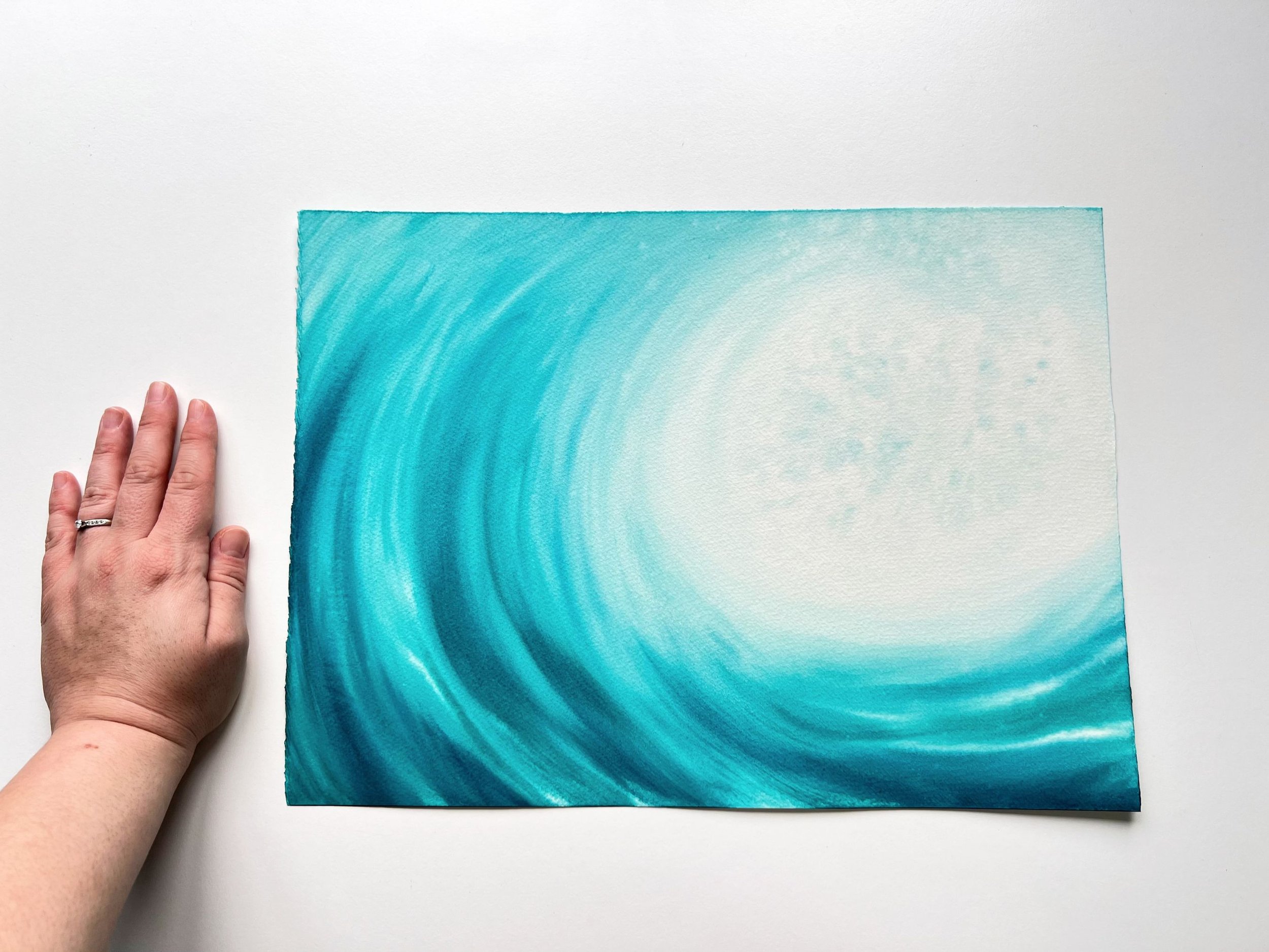 Closeup with my hand for size of the minimal watercolor painting "Blue Serenity" featuring a crashing wave with light blue hues. Ideal for coastal-inspired decor.