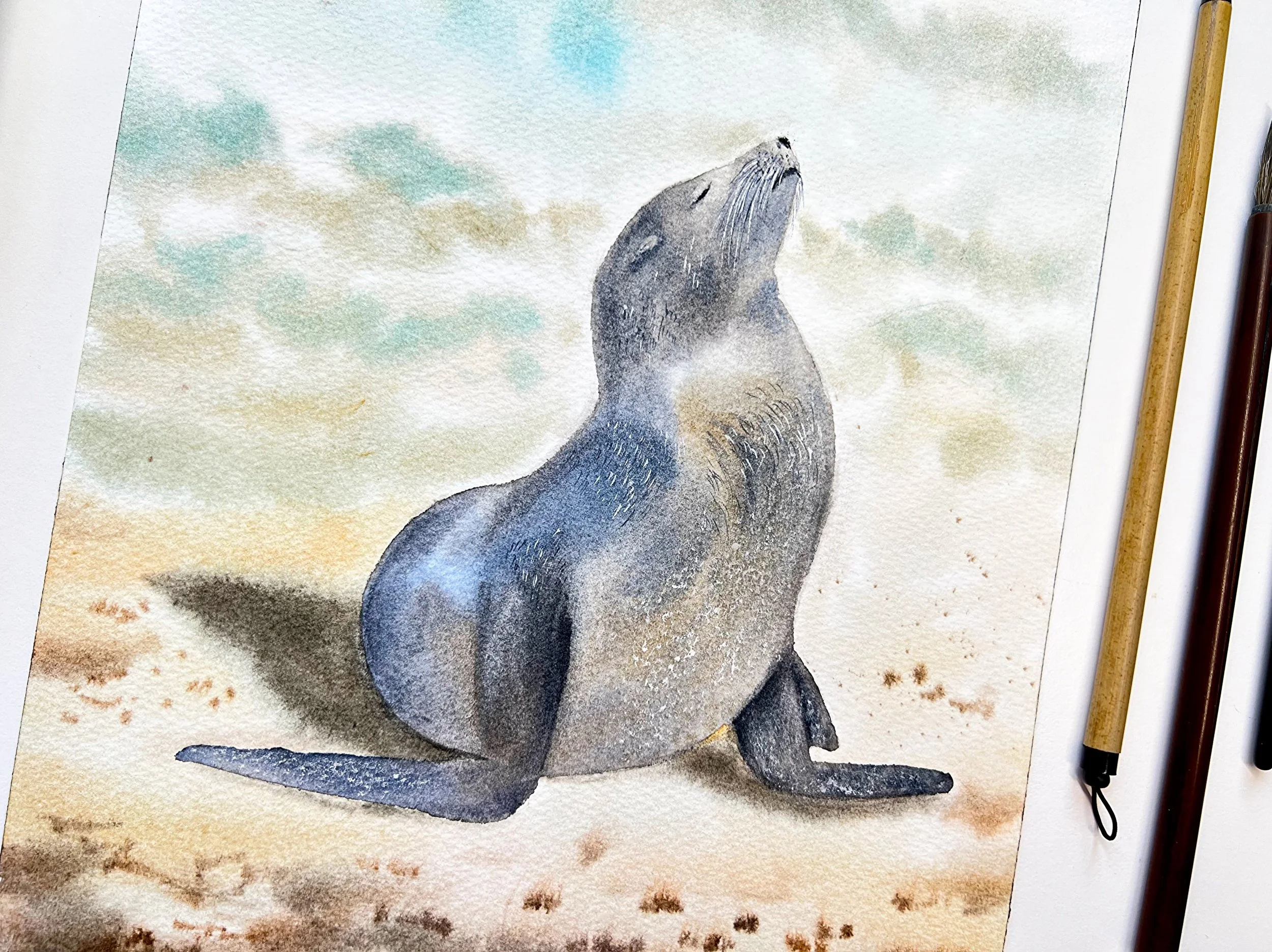 Close up of an original watercolor features a gentle Sunbathing Seal on the beach, painted in impressionistic style. in a setting with the original art supplies