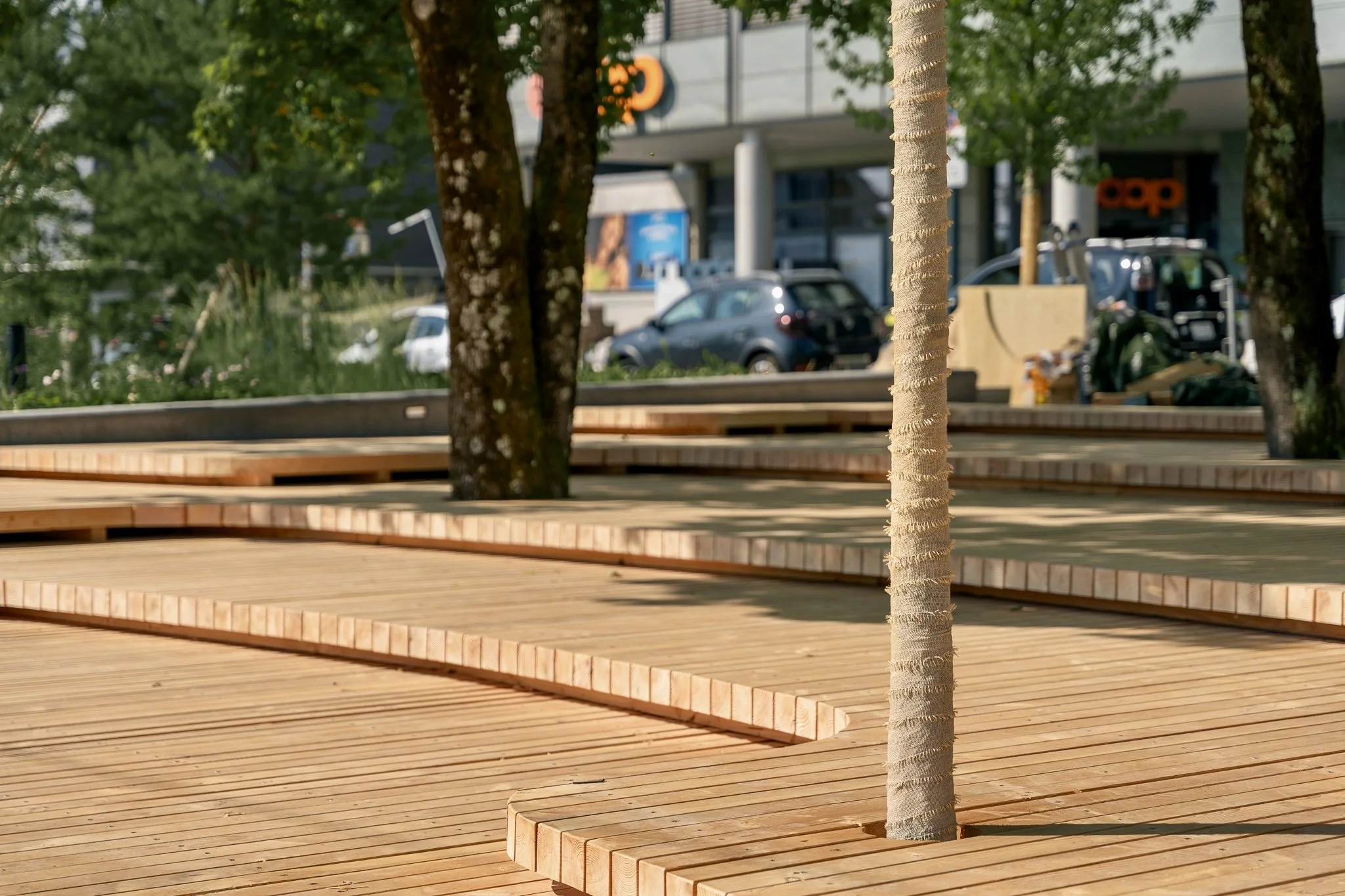 Holzterrasse mit mehreren Ebenen, umgeben von Bäumen, im Hintergrund ist eine moderne Gebäudefassade mit Fenstern und Parkplätzen.
