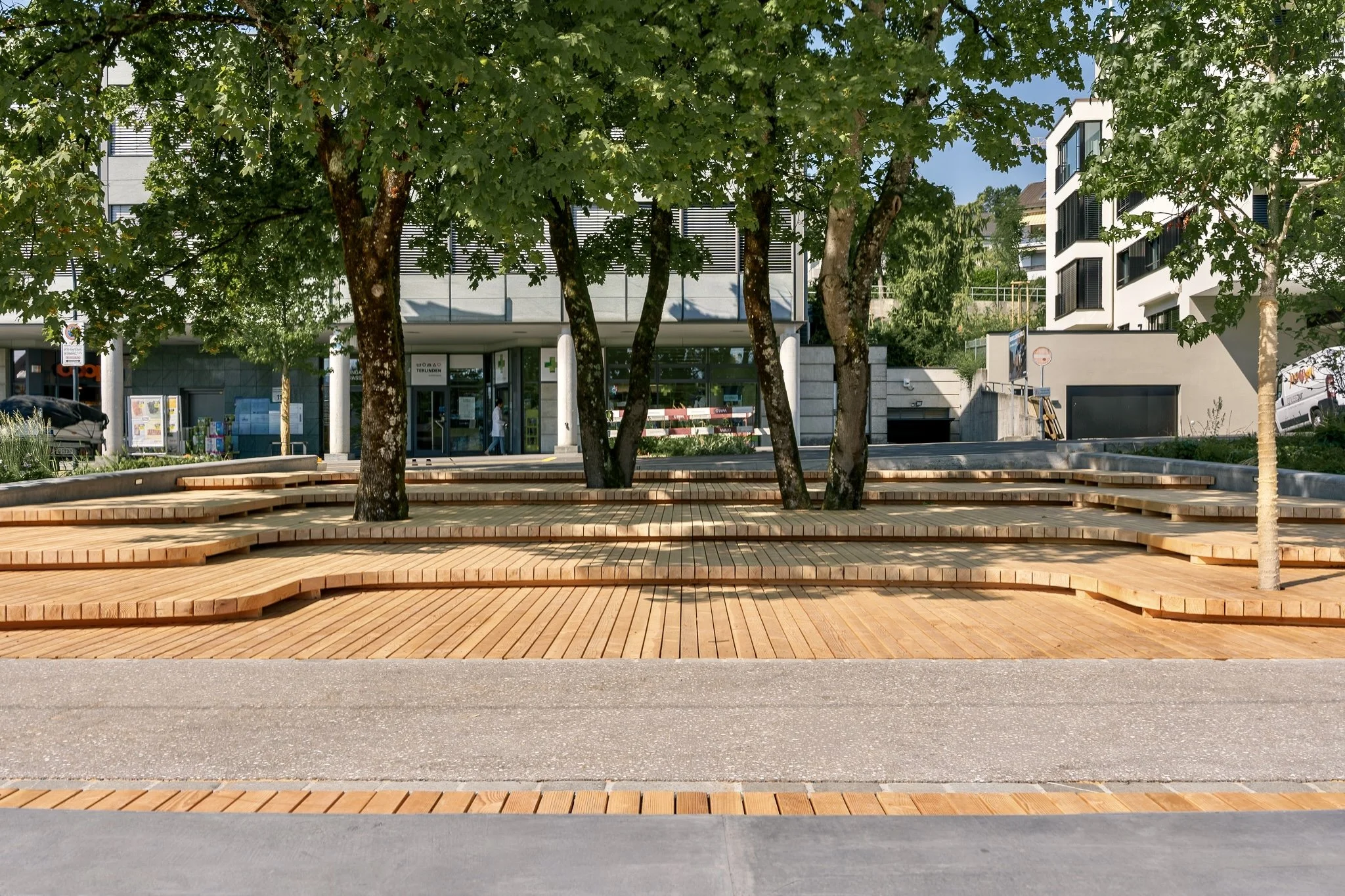 Ein öffentlicher Platz mit Bäumen und modernen Gebäuden im Hintergrund, gepflastertem Boden und Sitzbänken.