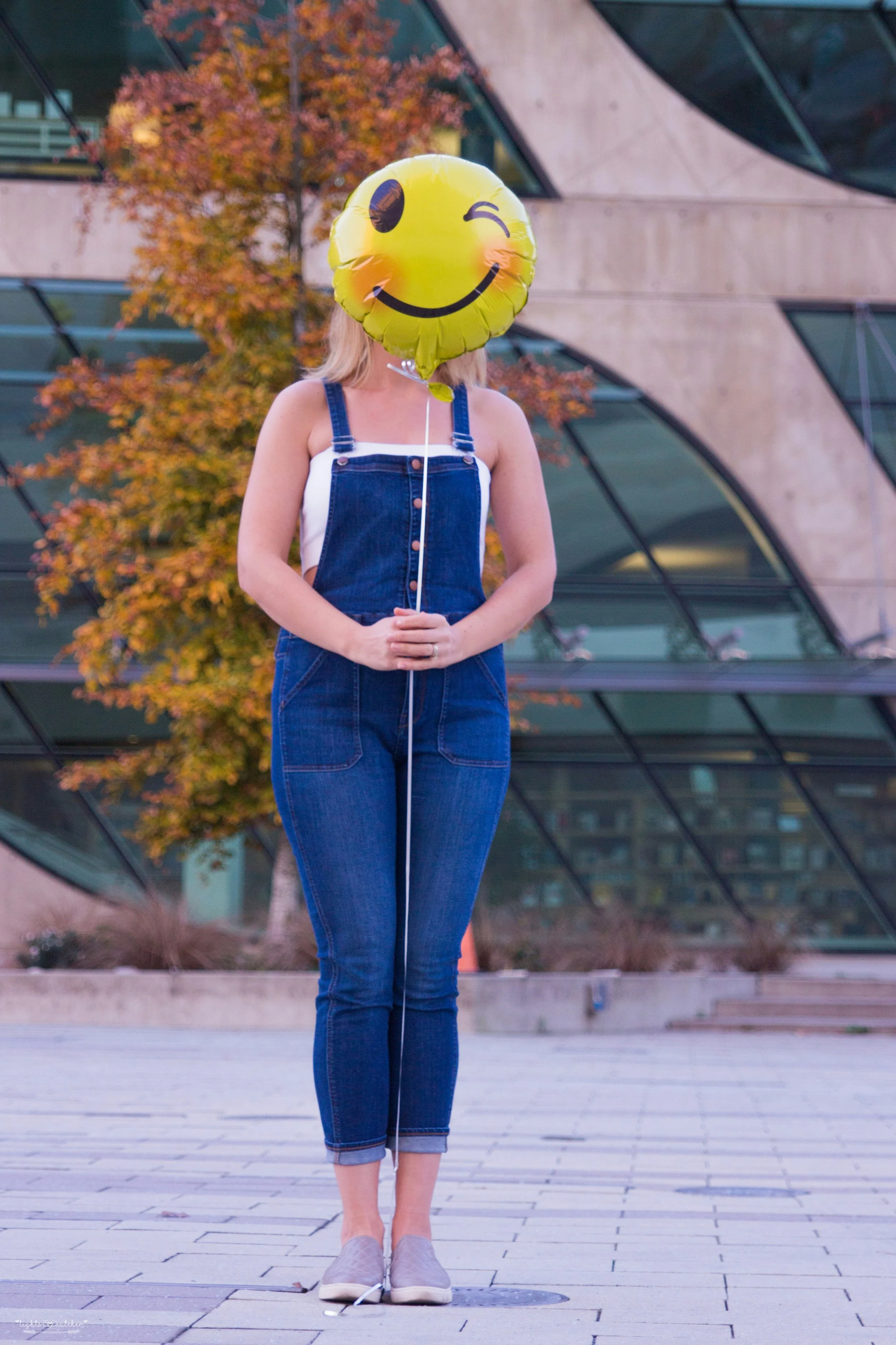 woman in dungarees whose face is obscured by a yellow 'smiley face' balloon
