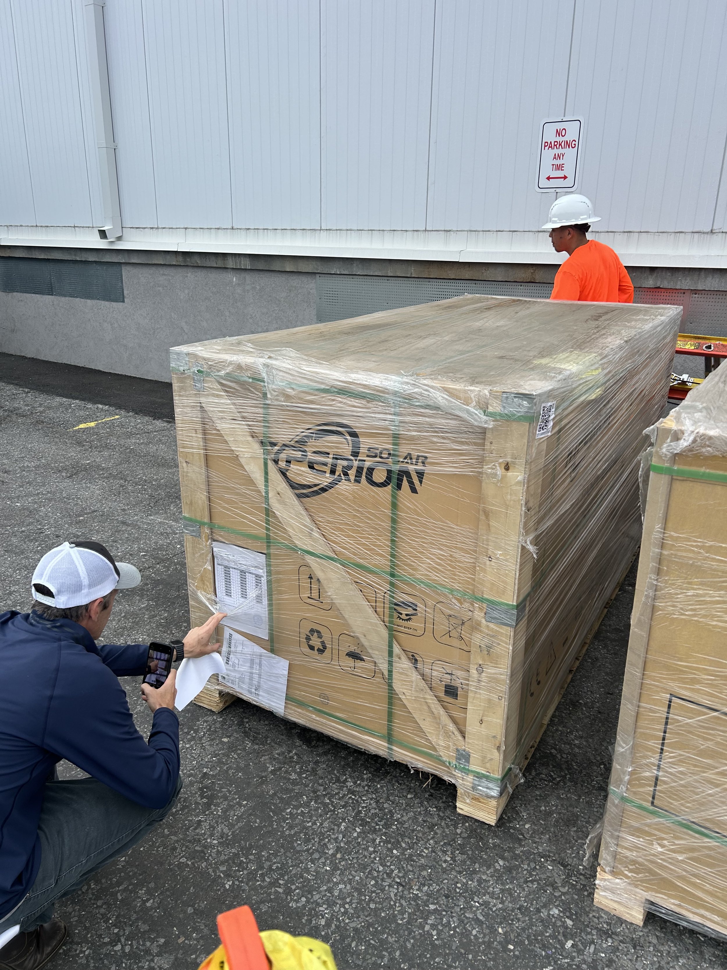 Man kneeling and taking a photo of a large wrapped pallet, with a worker in an orange shirt and white helmet standing nearby, at an outdoor loading area with a 'No Parking' sign on the wall.