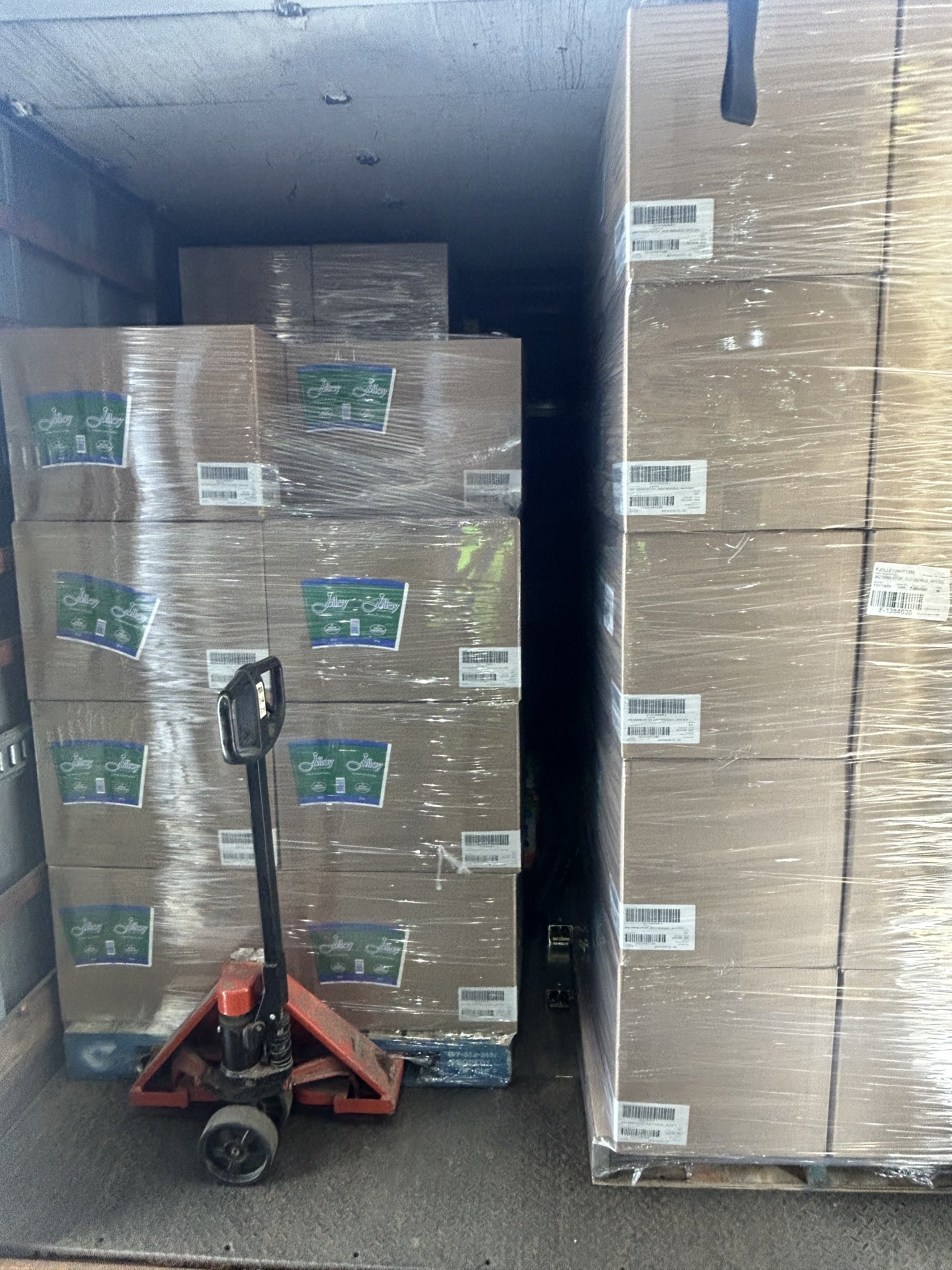 Stack of large boxed products wrapped in plastic, located inside a delivery truck, with a red pallet jack in front of the boxes.
