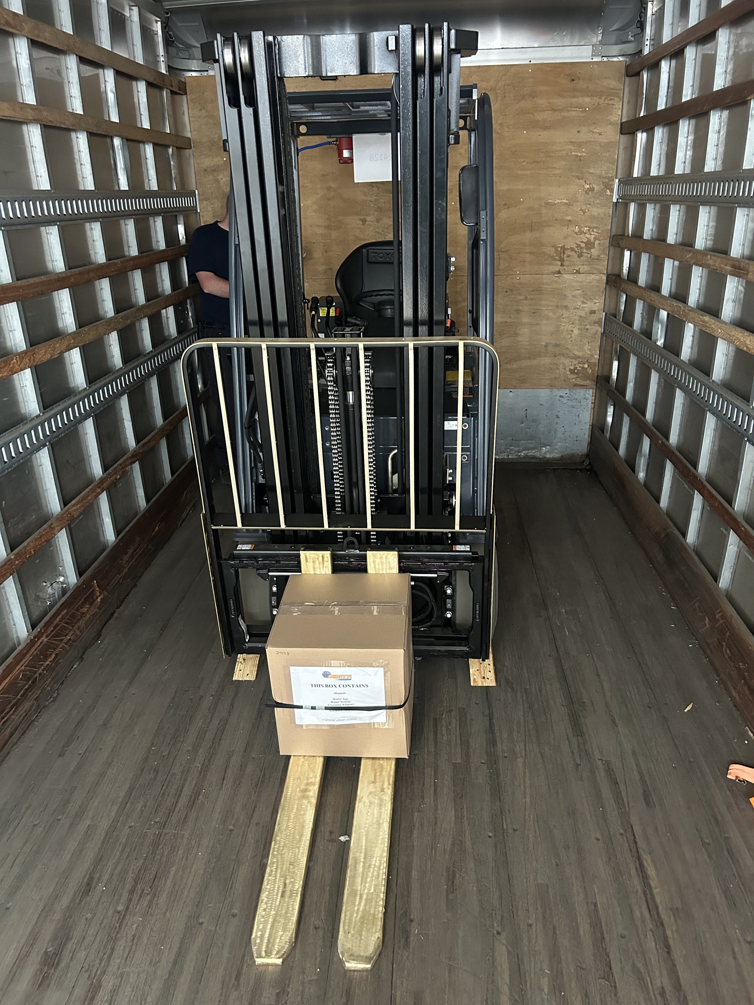 A forklift inside a moving truck, with a cardboard box on its forks, ready for transportation.