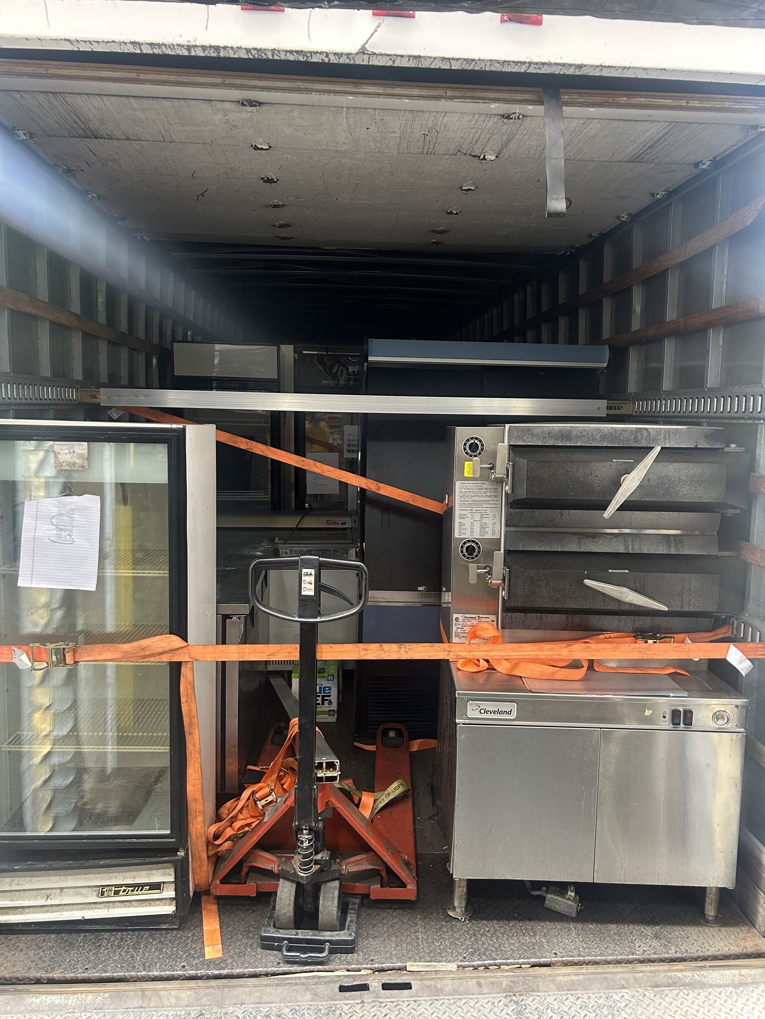Inside a moving truck, there are commercial kitchen appliances including a small refrigerator, a pizza oven, and a stainless steel freezer, all secured with orange straps.