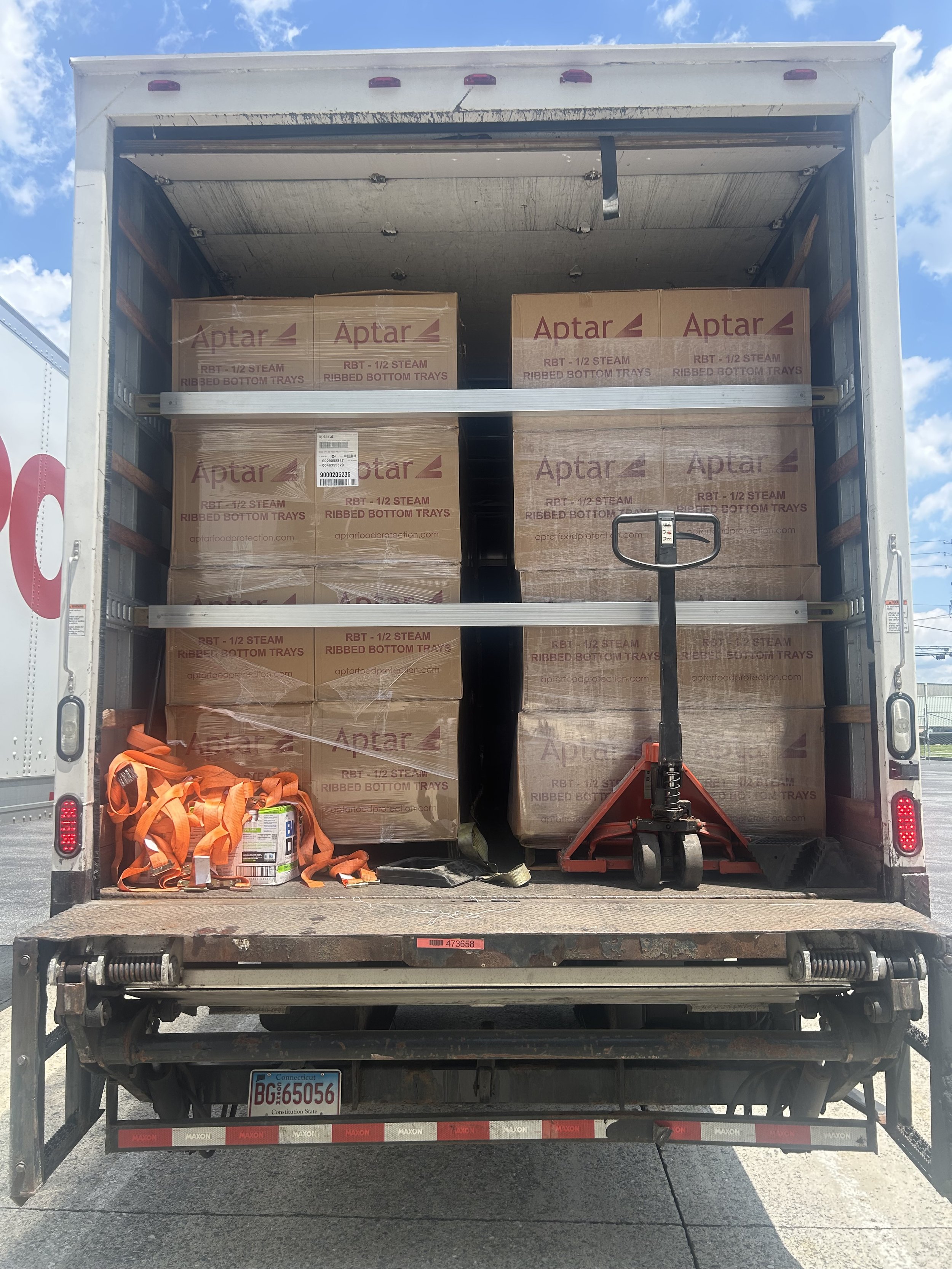 Back of a delivery truck filled with boxes labeled Aptar, containing ribbed bottom trays for food transport, and some orange safety straps and a hand truck in the truck bed.