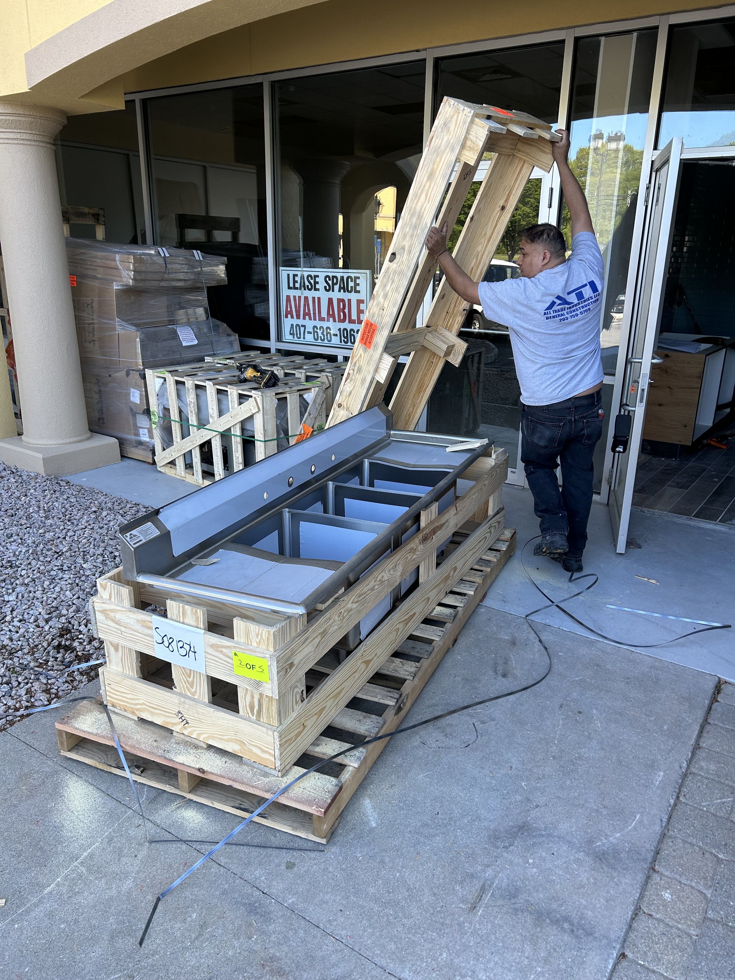 Man assembling a wooden frame outside a store with a sign that reads 'Lease Space Available'