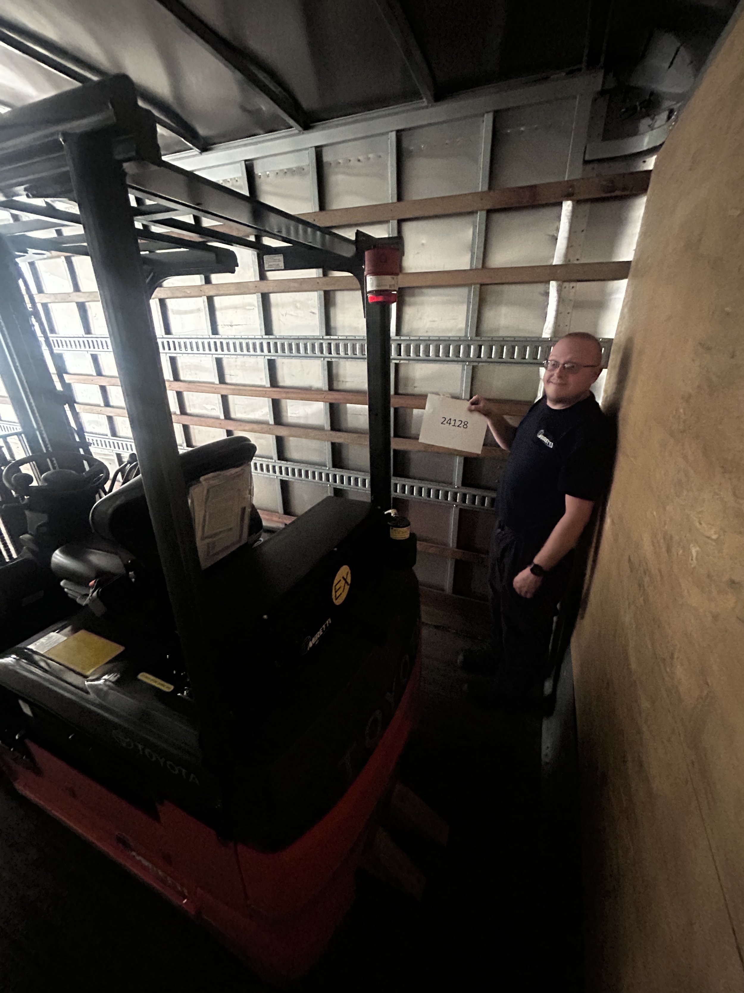 Man standing next to a forklift inside a storage unit, holding a sign with the number 24128.